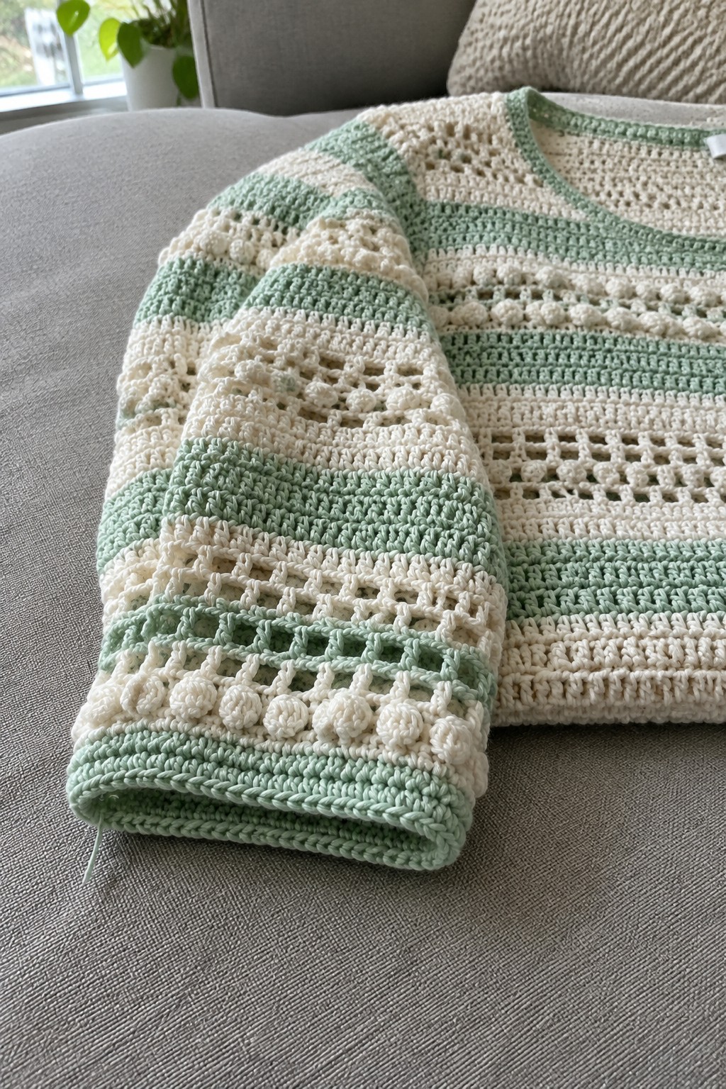 Striped mint green and cream crocheted sweater with bobble and lace patterns on gray couch.