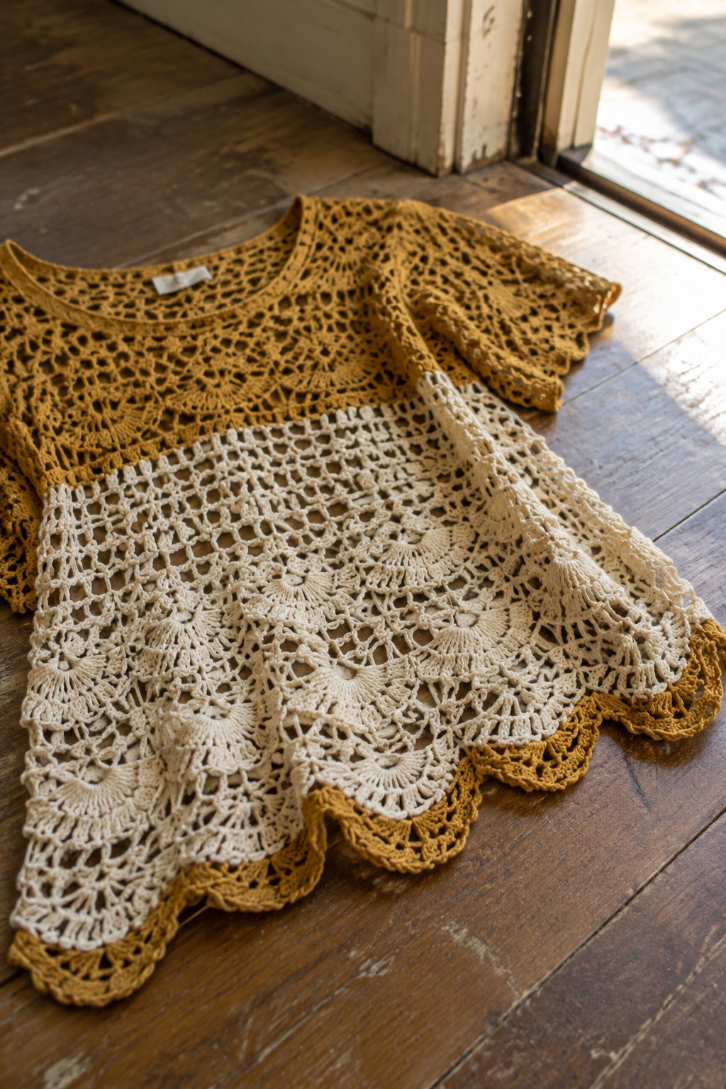 A two-tone crocheted lace top in mustard and cream laid flat on wood flooring, displaying openwork patterns and a scalloped hem.