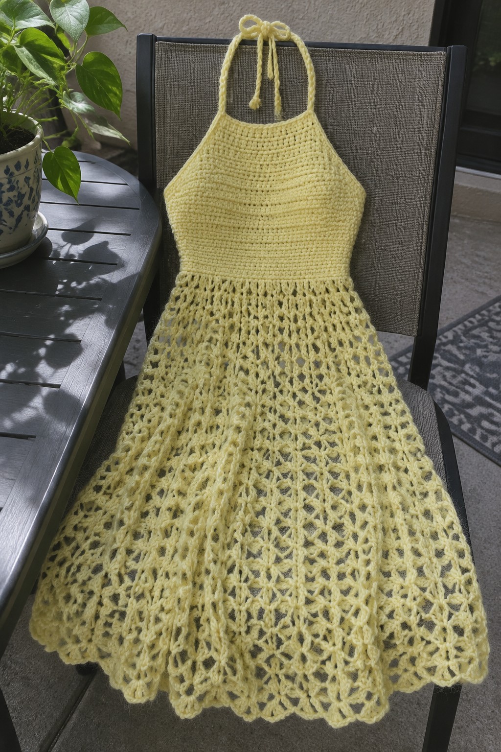 A pale yellow crocheted halter dress with a solid bodice and lacy flared skirt hangs on a black chair outdoors.