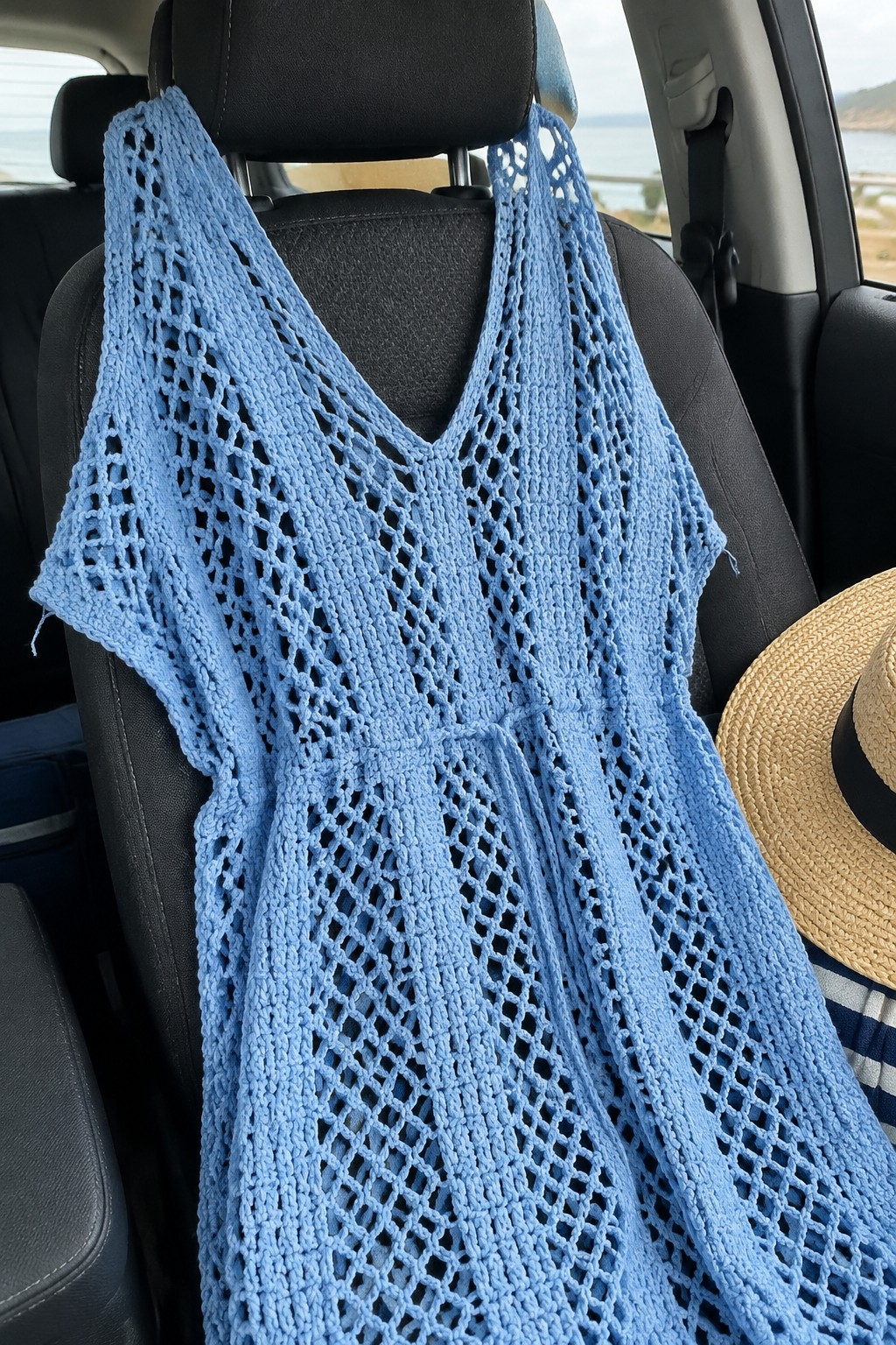 Light blue open-mesh crochet dress draped over a black car seat with a straw hat visible on the right.