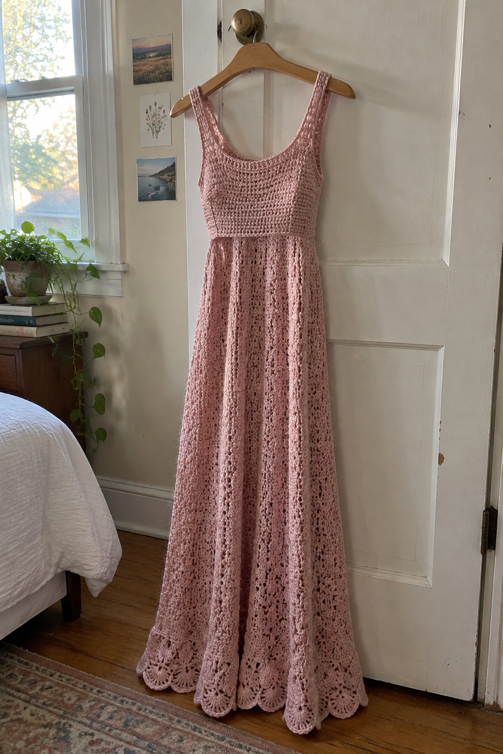 Pink crocheted maxi dress with scalloped hem on wooden hanger against white door.