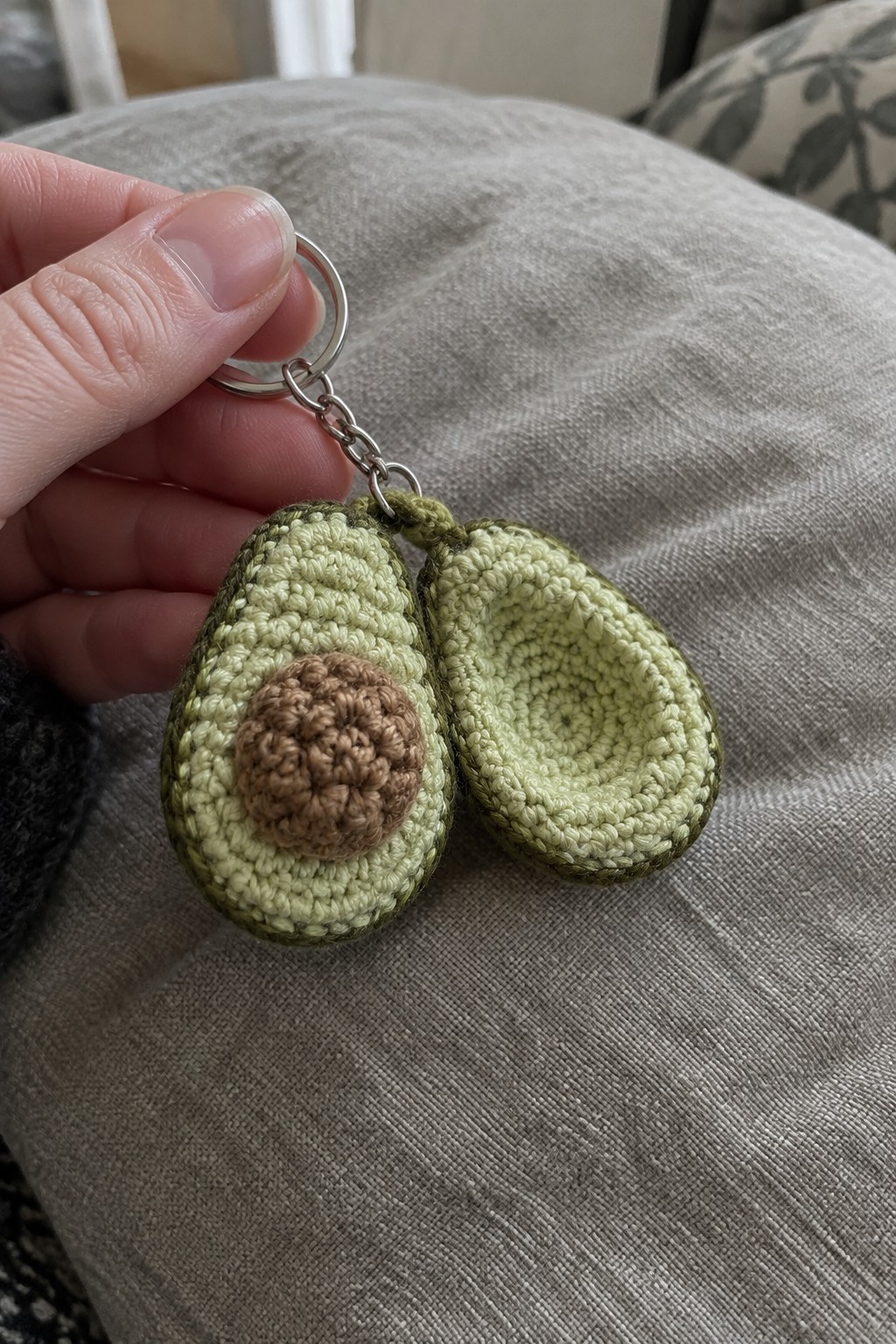 Hand holding crocheted avocado keychain with two green halves and brown pit