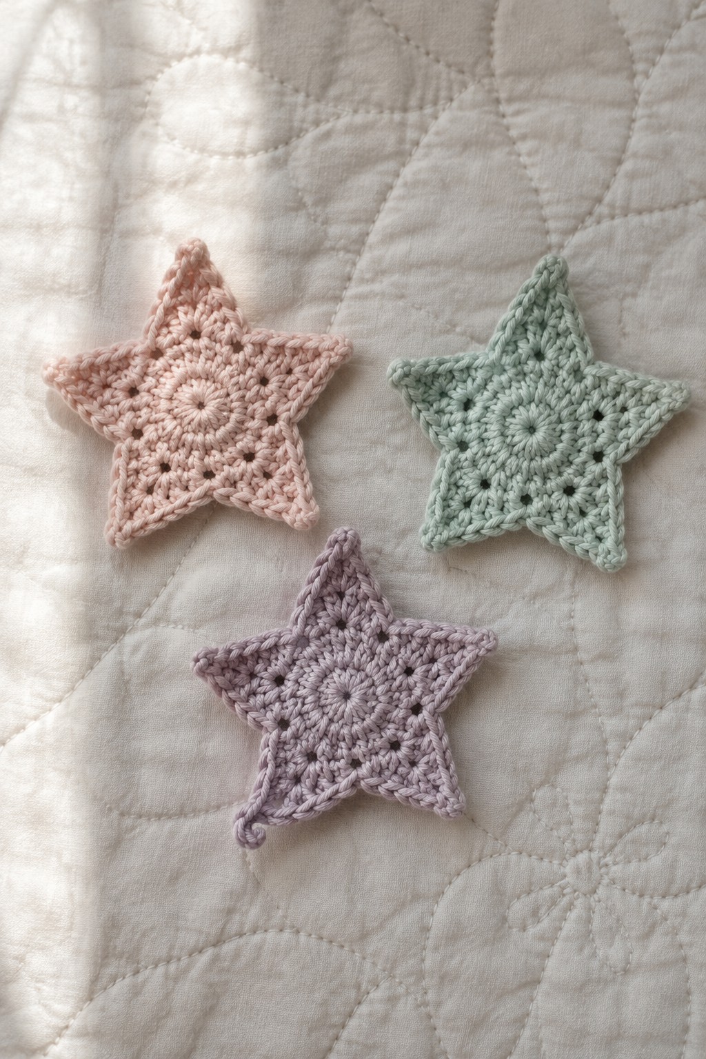 Three pastel crocheted stars on a white quilted fabric in soft sunlight.