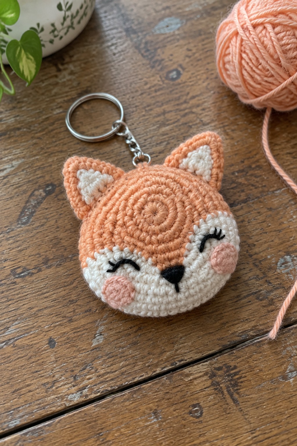 Crocheted orange fox keychain with closed eyes on wooden table beside yarn ball
