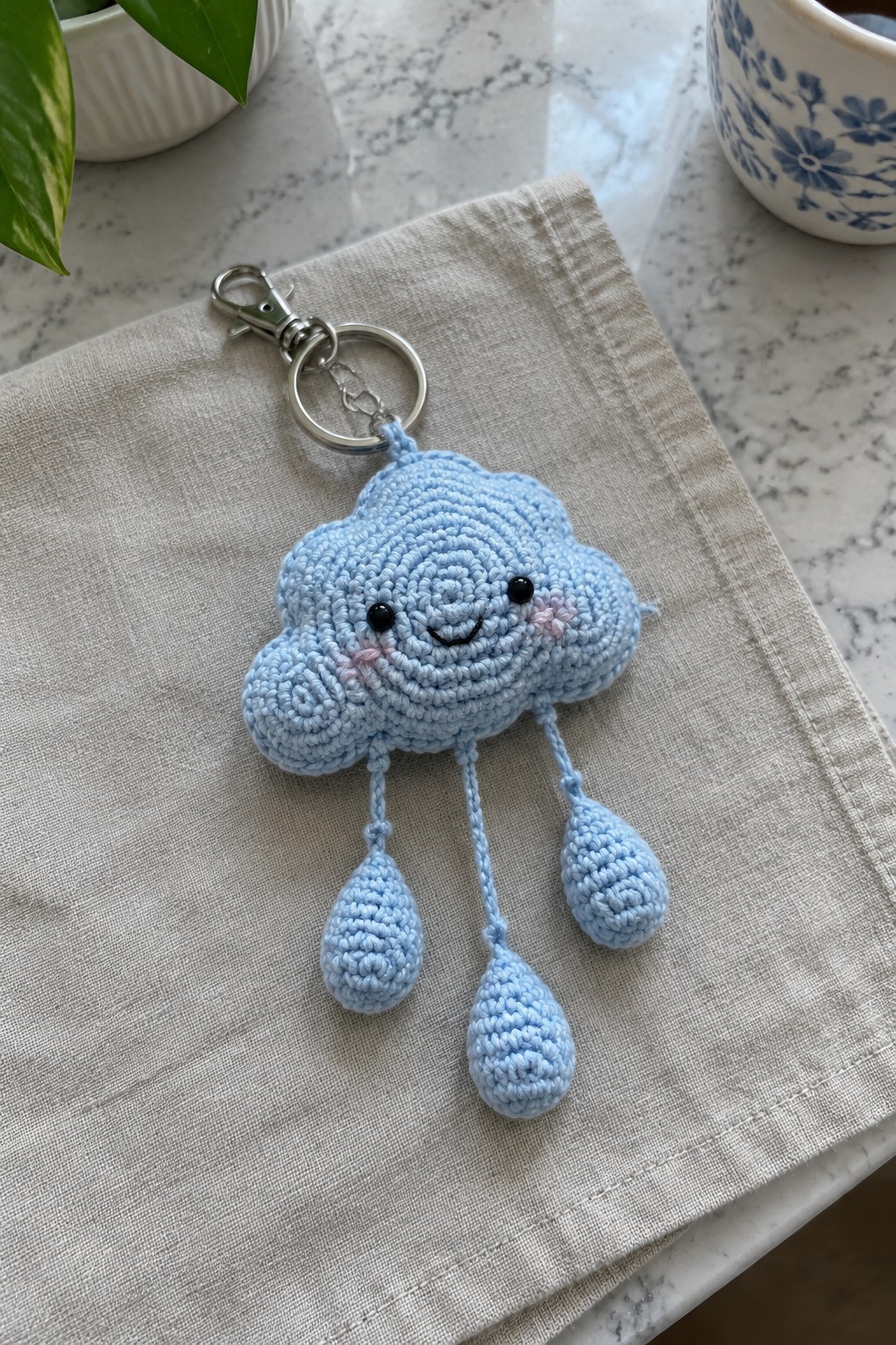 Light blue crocheted cloud keychain with smiling face and dangling raindrops on beige cloth