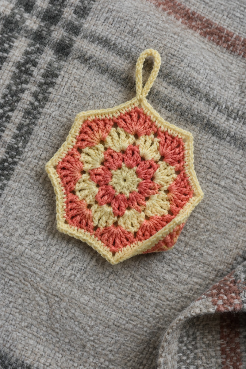 Crocheted hexagonal potholder with pink-yellow floral pattern and hanging loop on woven fabric