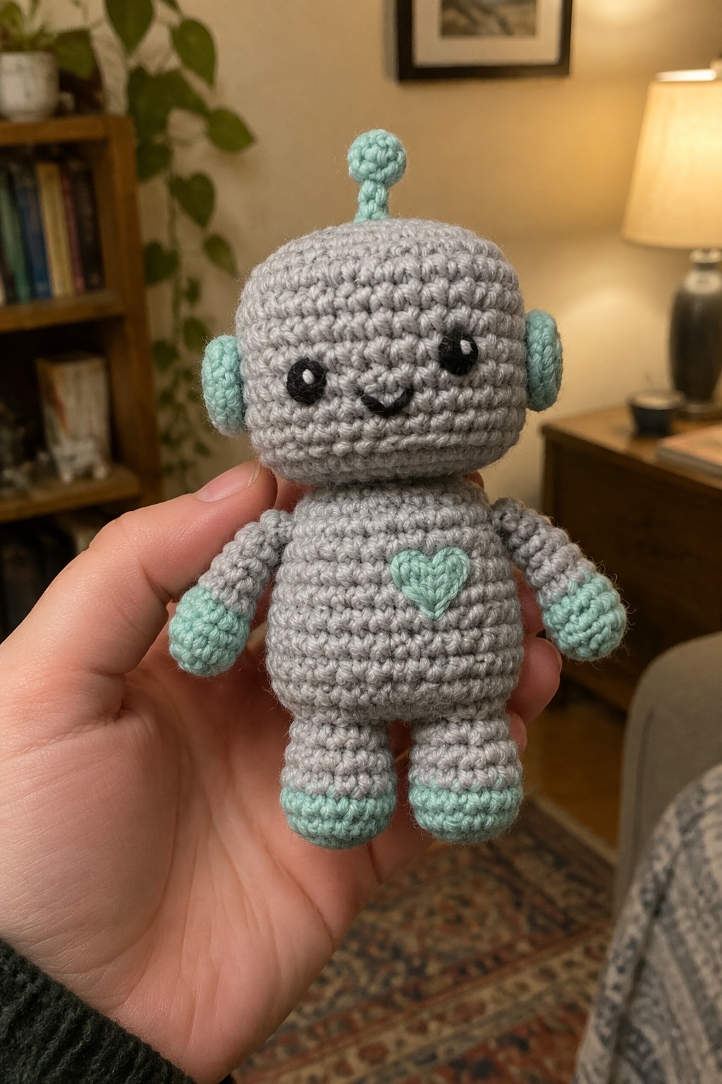 Hand holding gray crocheted robot with mint green heart, antenna, and limbs
