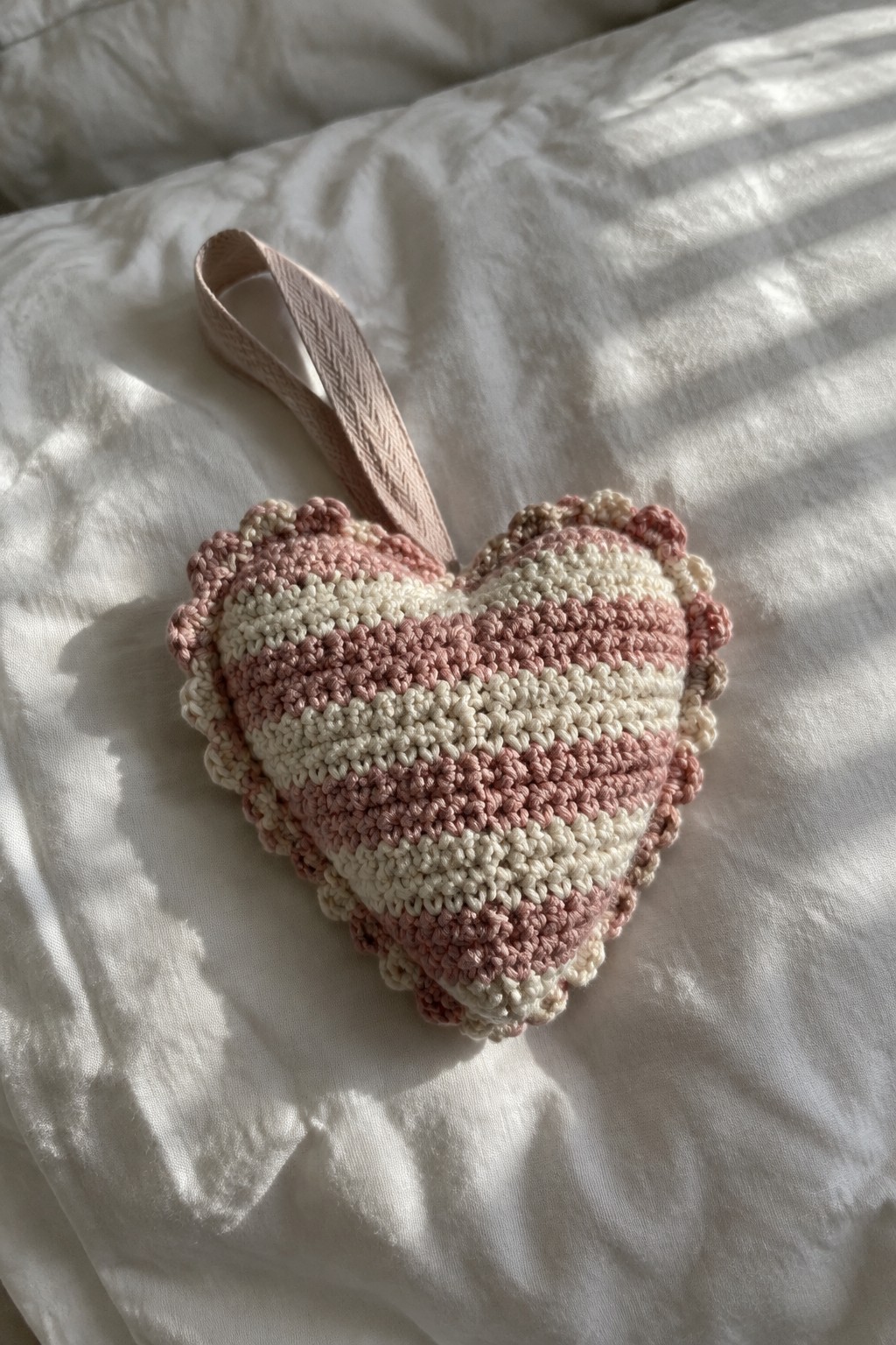 Crocheted pink and white striped heart pillow with ribbon loop on white bedding