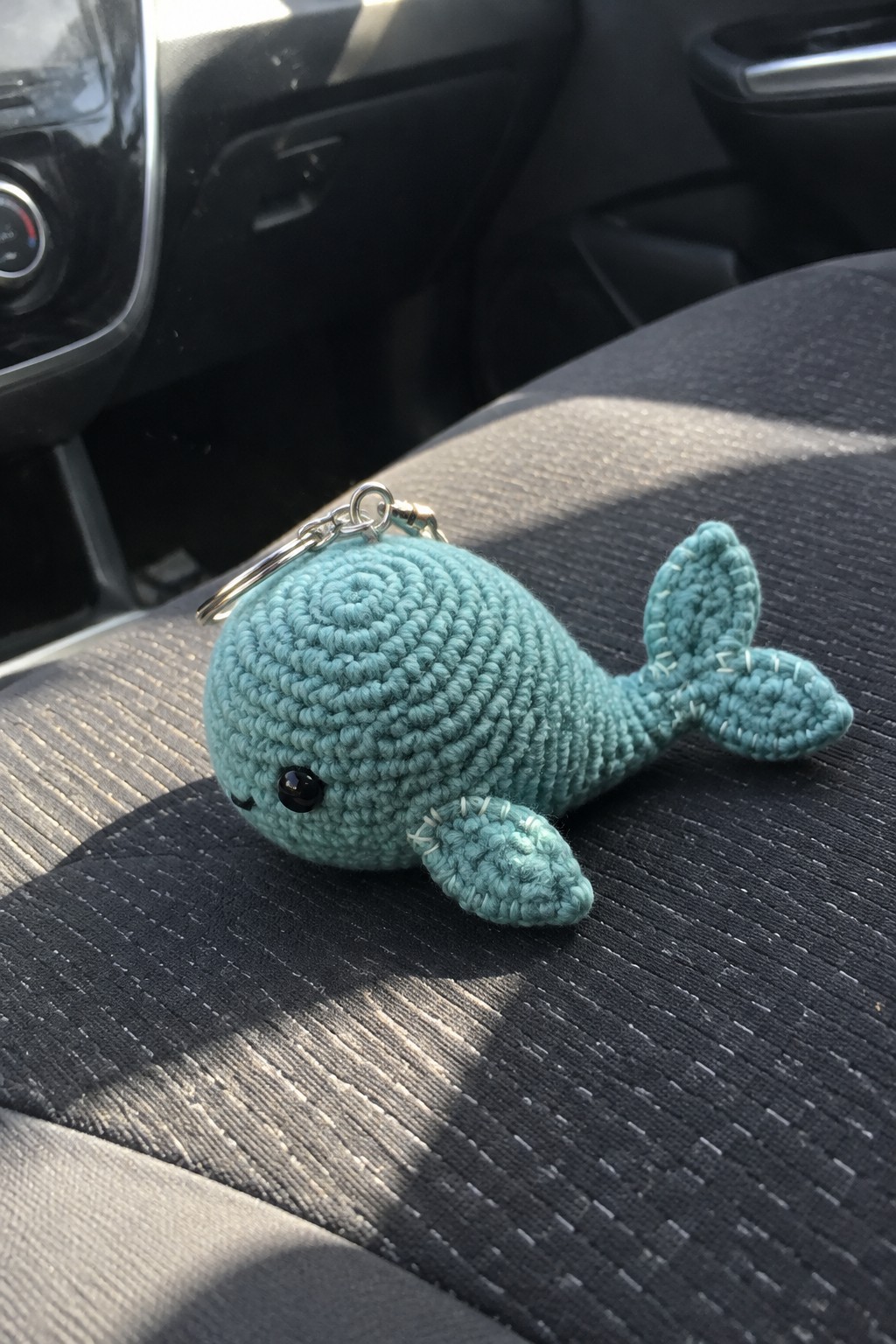 Teal crocheted whale keychain with ring on gray car seat in sunlight.