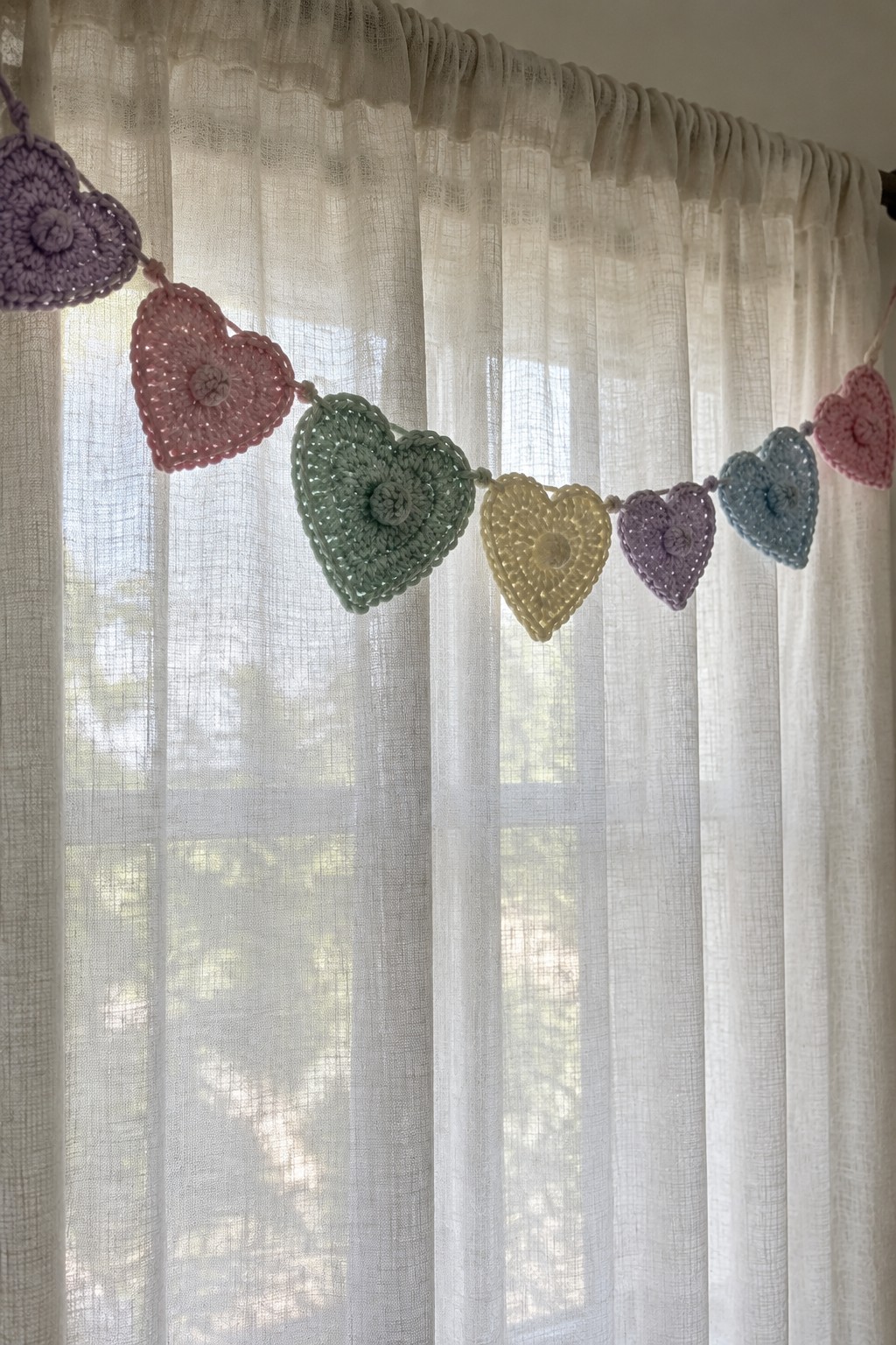 Colorful crocheted heart garland hanging before sheer white window curtains.