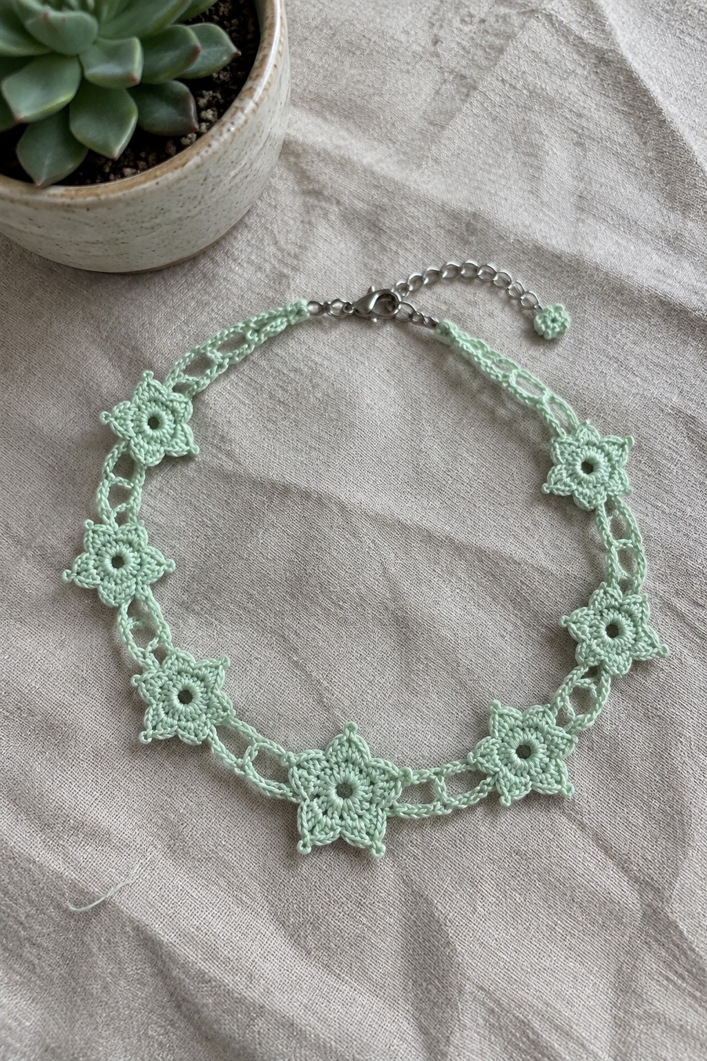 A mint green crocheted necklace with multiple flower motifs linked by open chains, laid on beige fabric beside a potted succulent.