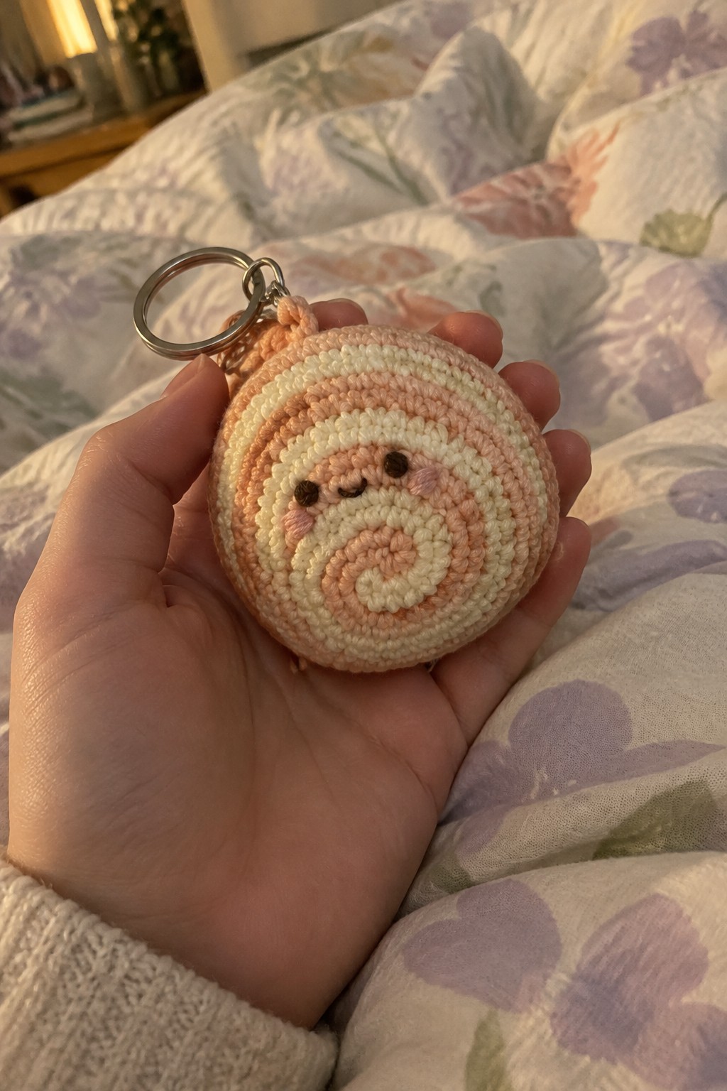 Hand holding pastel striped crochet snail keychain with metal ring on floral bedding