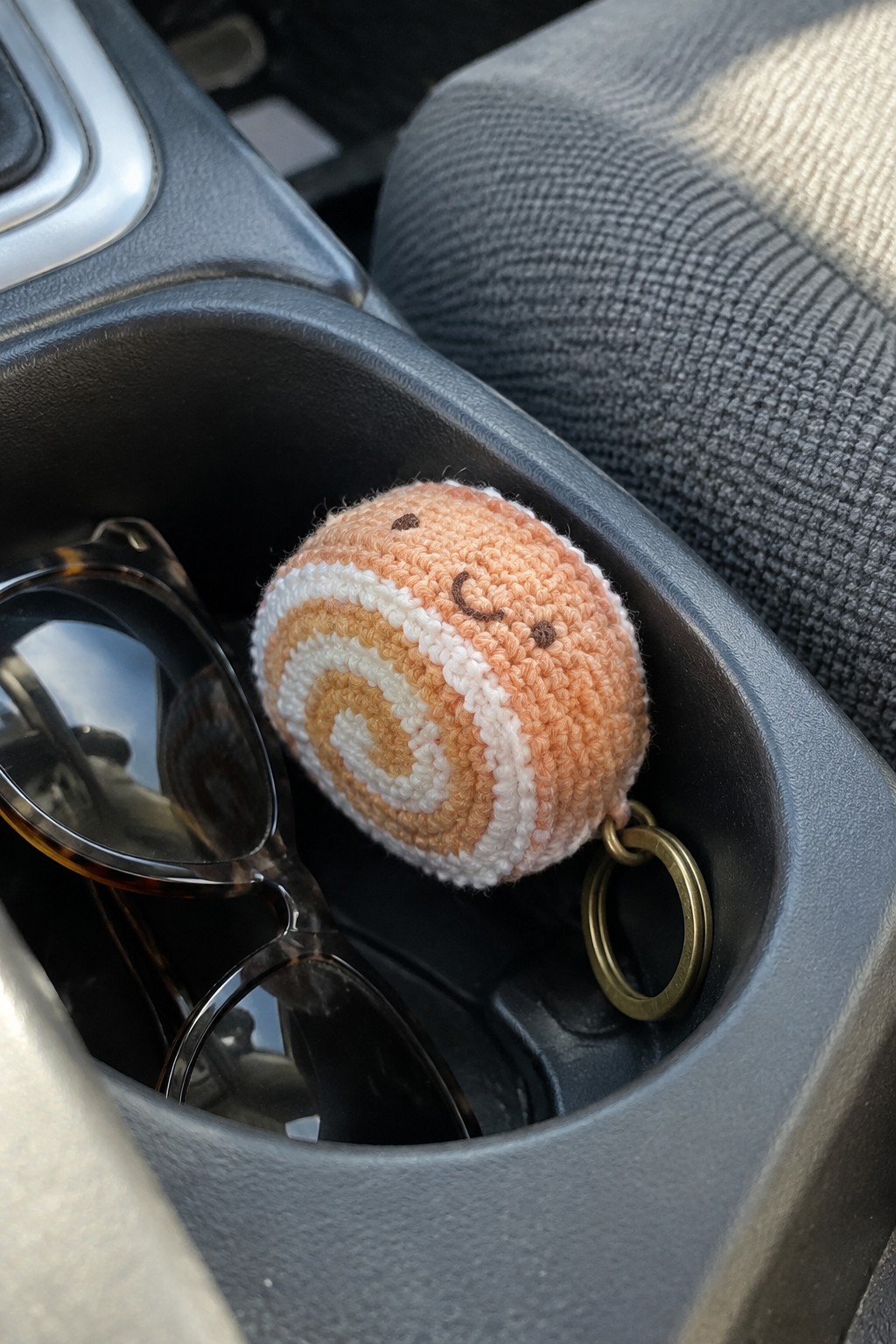 Crocheted cinnamon roll keychain with face in car cup holder beside sunglasses
