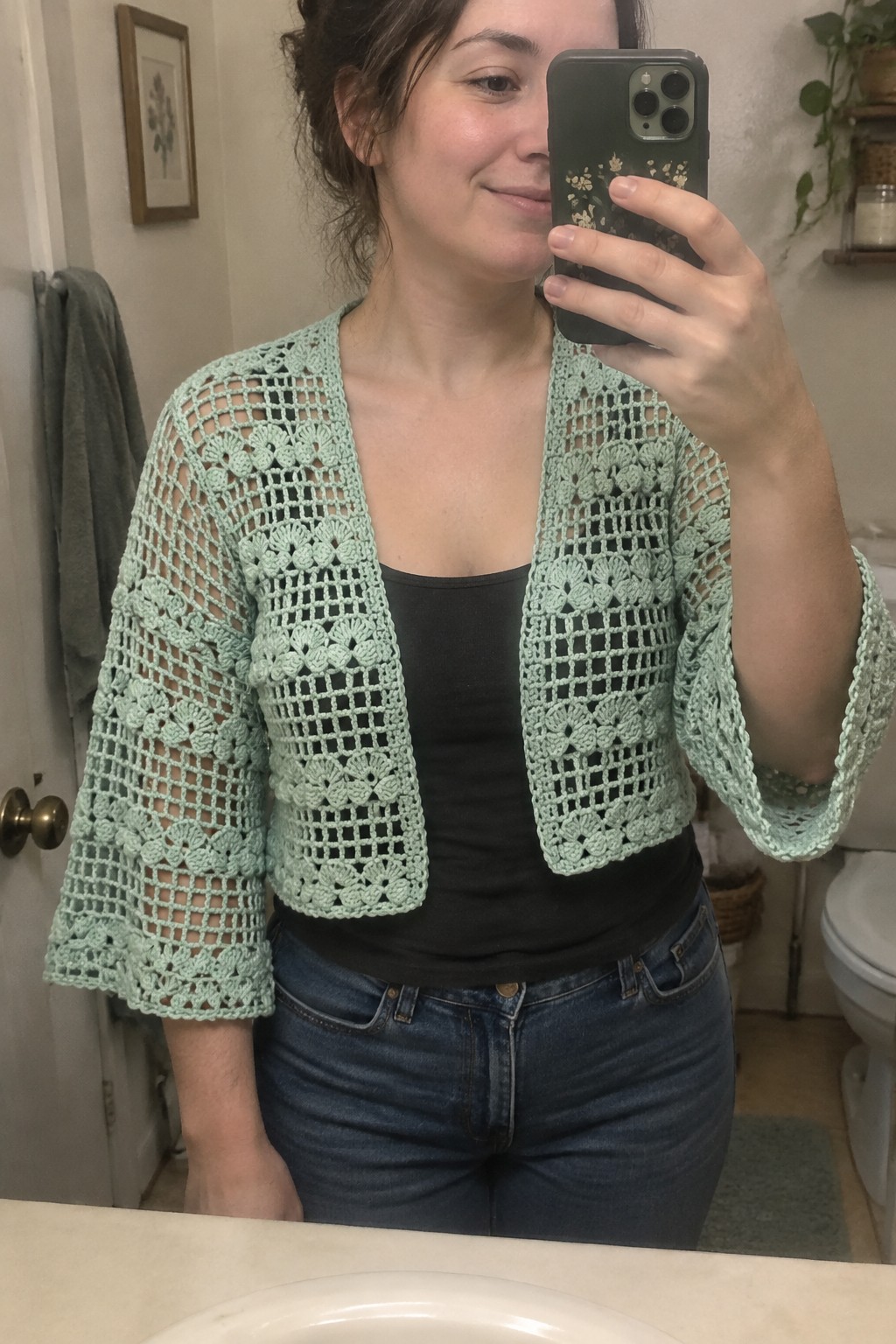 Woman in mint green crochet cardigan taking mirror selfie in bathroom wearing jeans