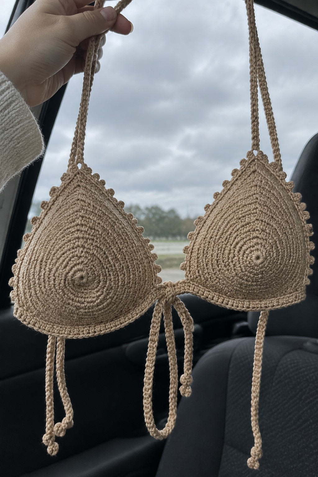 A hand holds a beige crocheted bikini top with spiral cups and long ties inside a car.