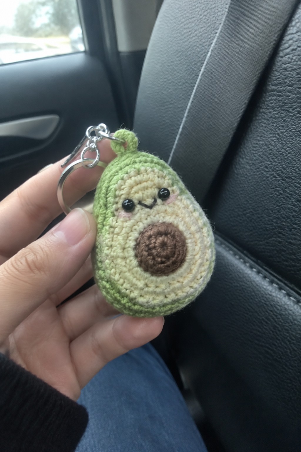 Hand holding green crocheted avocado keychain with cute face in car interior
