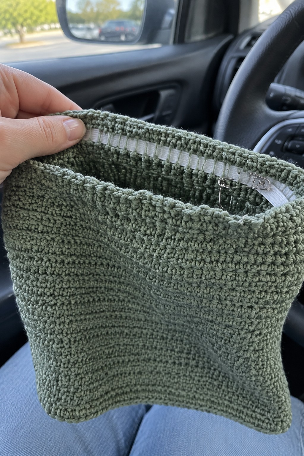 A hand holds open a sage green crocheted tube showing white elastic sewn inside the top rim with a safety pin attached.