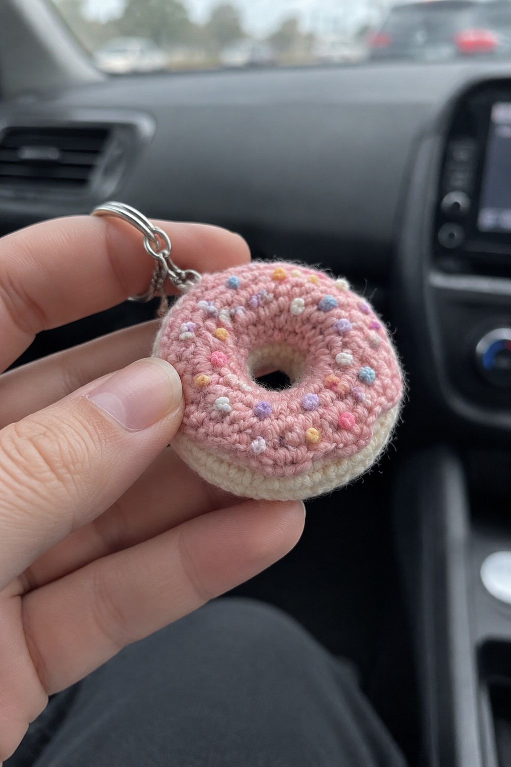 Hand holding pink crocheted donut keychain with colorful sprinkles in car interior