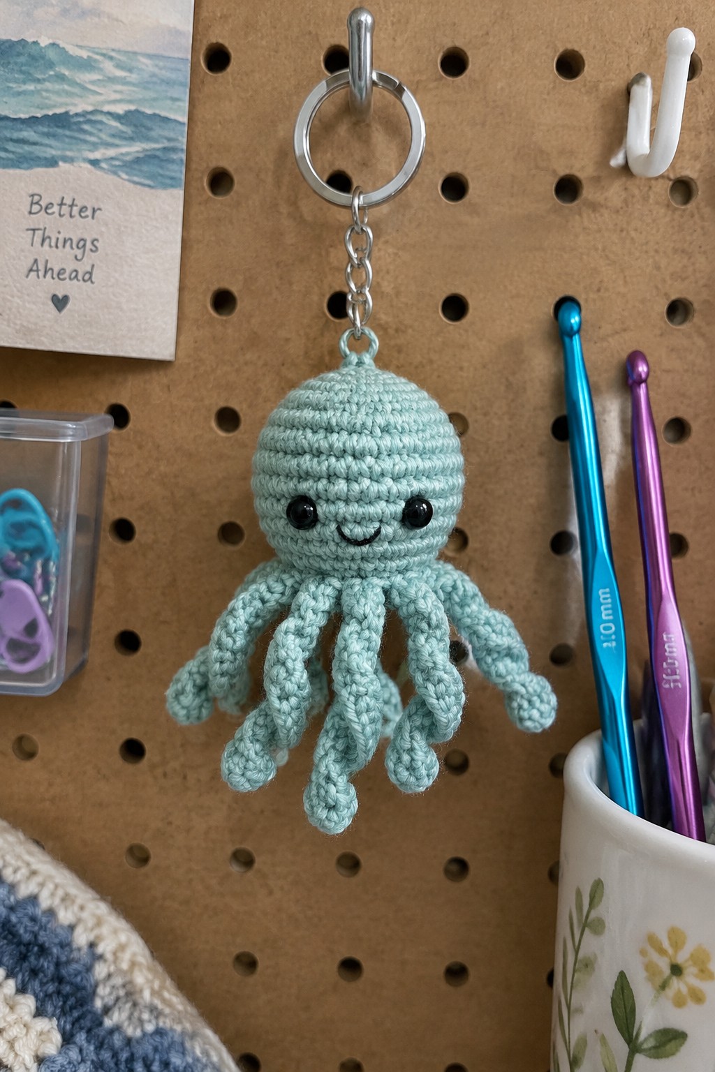 Crocheted mint green octopus keychain with smiling face on pegboard near crochet hooks
