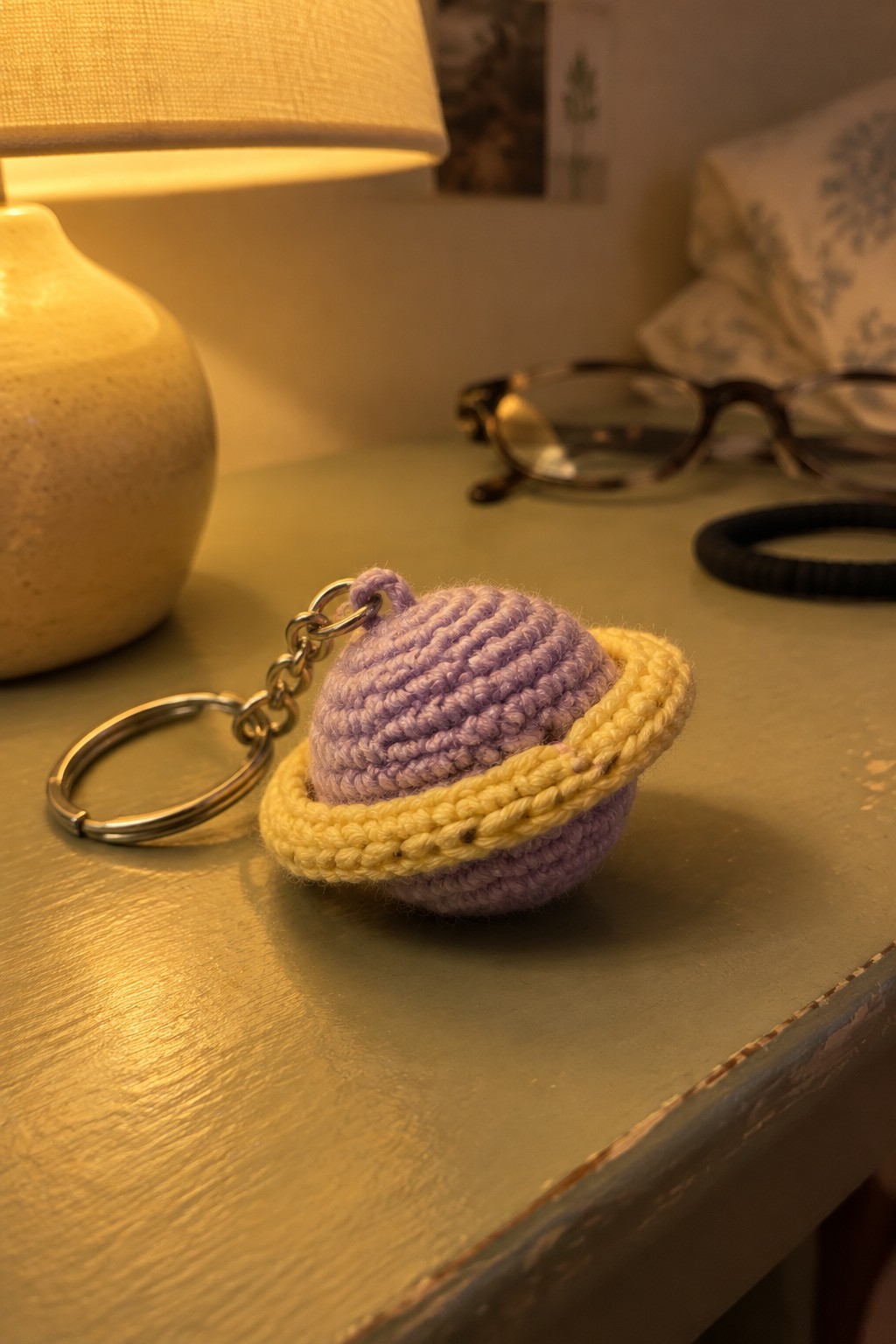 Crocheted purple planet keychain with yellow rings on green wooden table beside lamp.