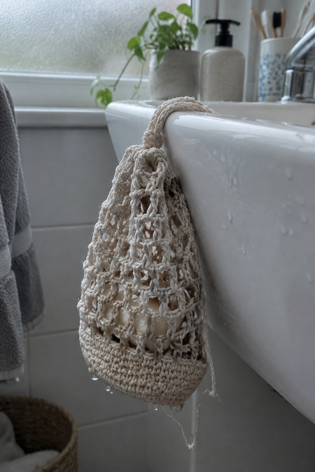 Beige crocheted mesh soap bag hanging from white bathroom sink with droplets