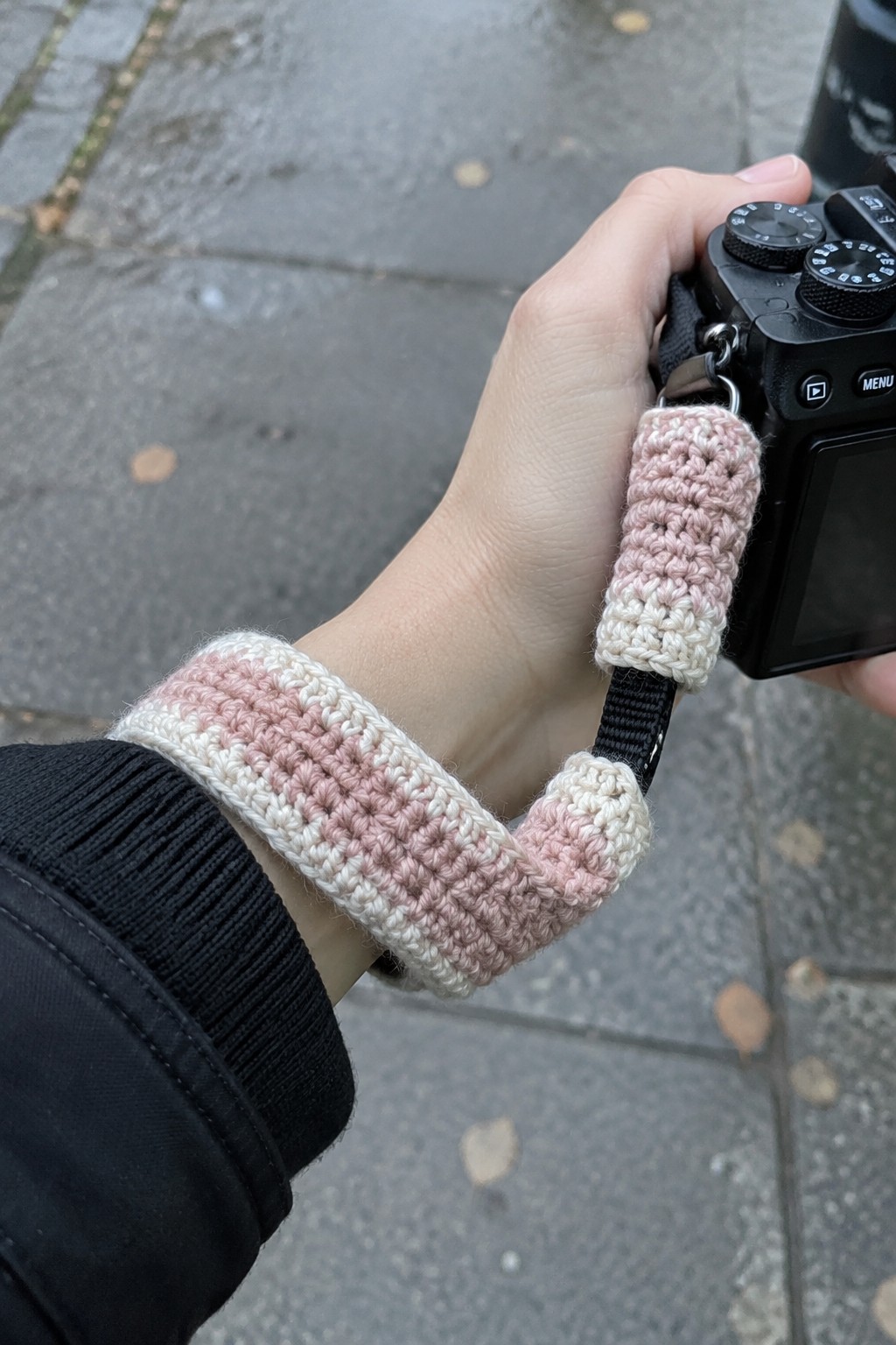 A person's wrist wearing a black jacket with a pink and cream striped crocheted cover on a camera strap attached to a black camera.