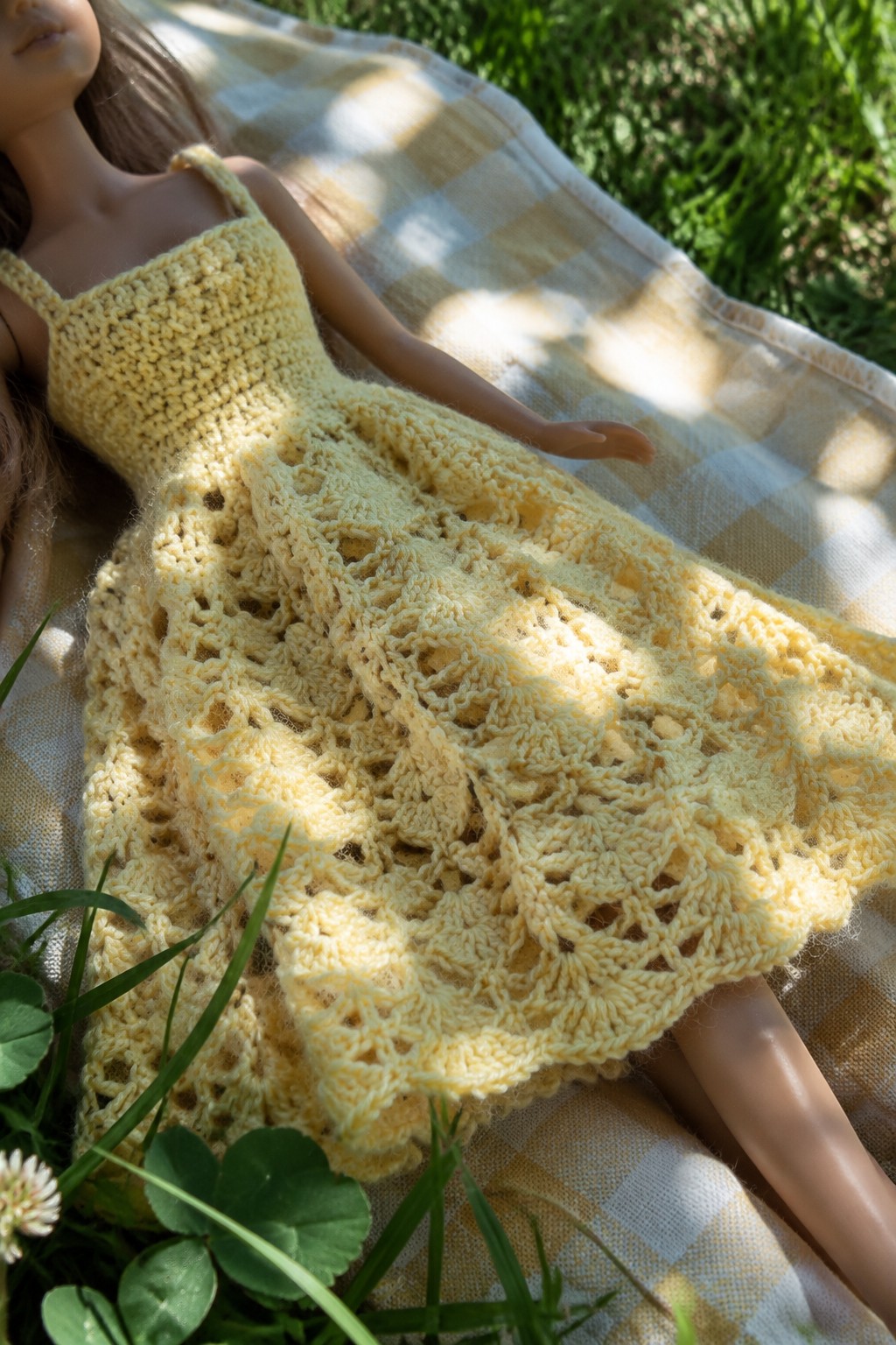 Doll in yellow crocheted lace dress reclining on checkered blanket in grass
