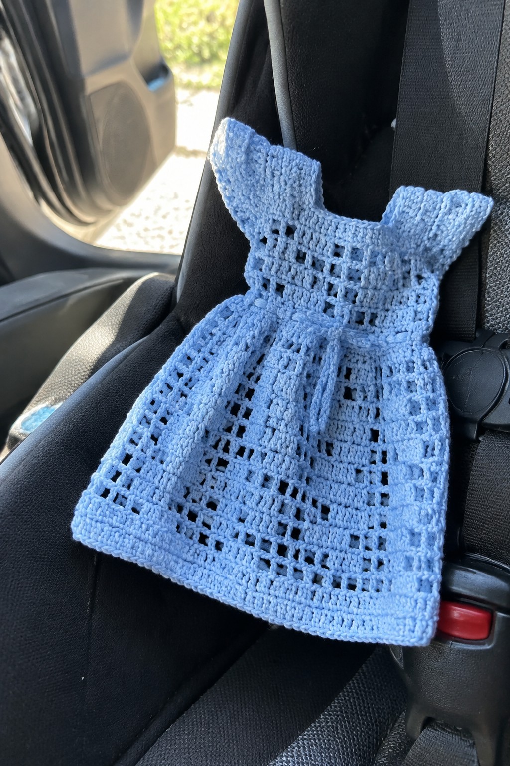 A light blue crocheted miniature dress with an openwork lace skirt and ruffled shoulders rests on a car seat next to a seatbelt.