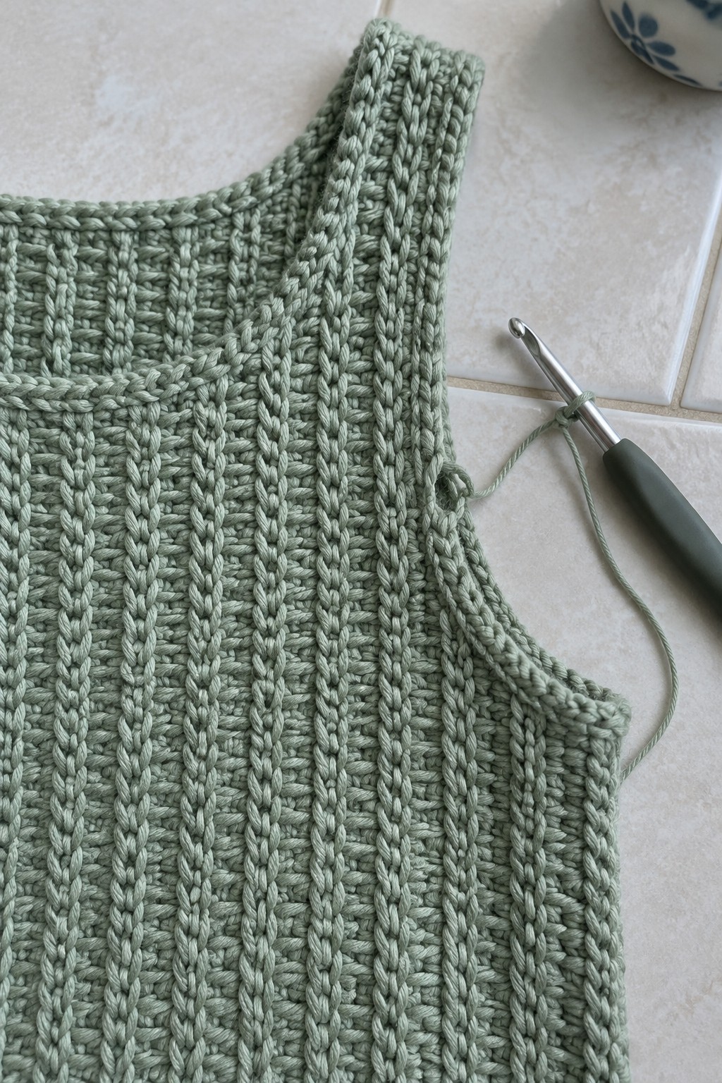 Light green ribbed crochet vest with hook and yarn on white tiles