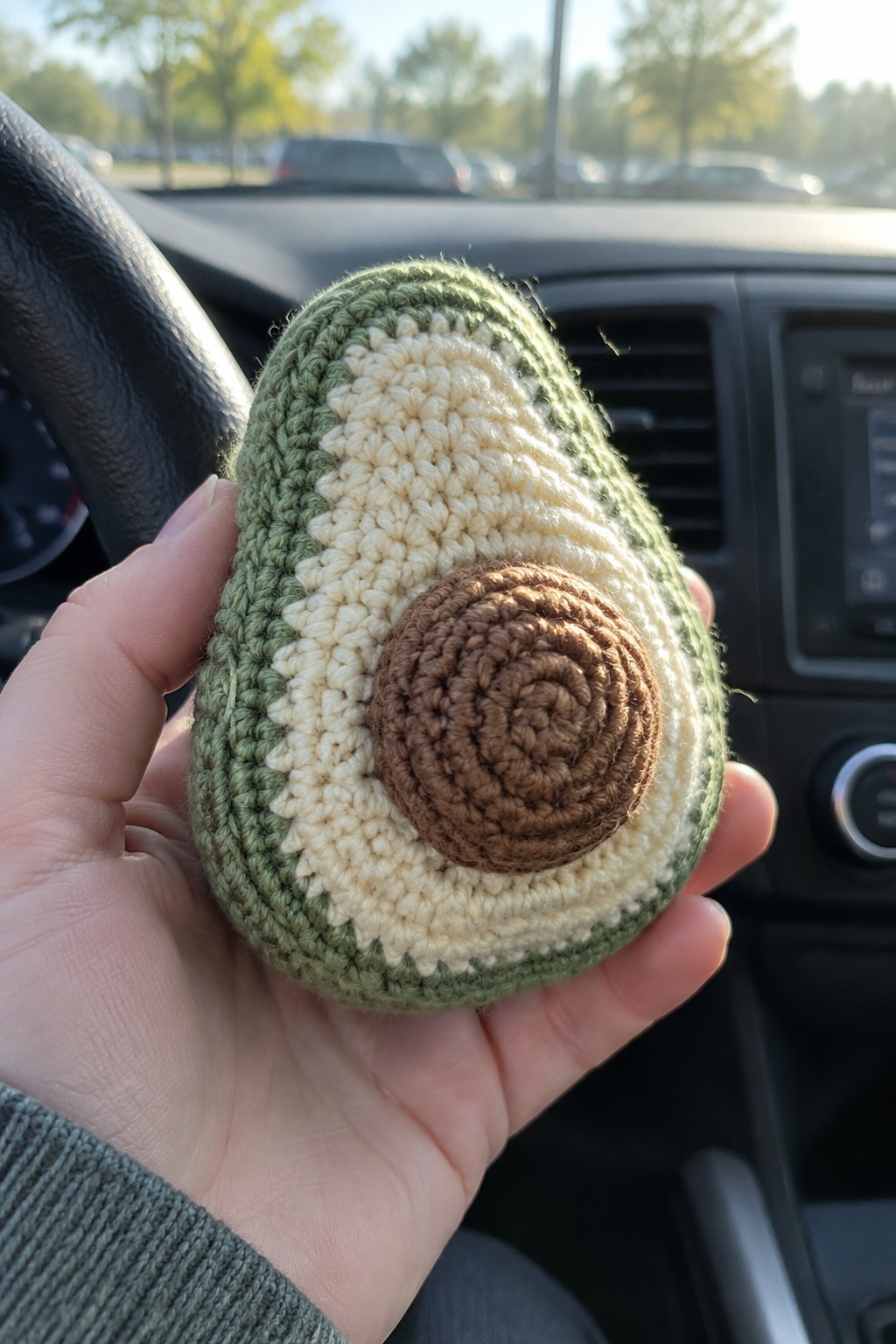 Hand holding crocheted avocado plush with green, beige, and brown yarn in a car