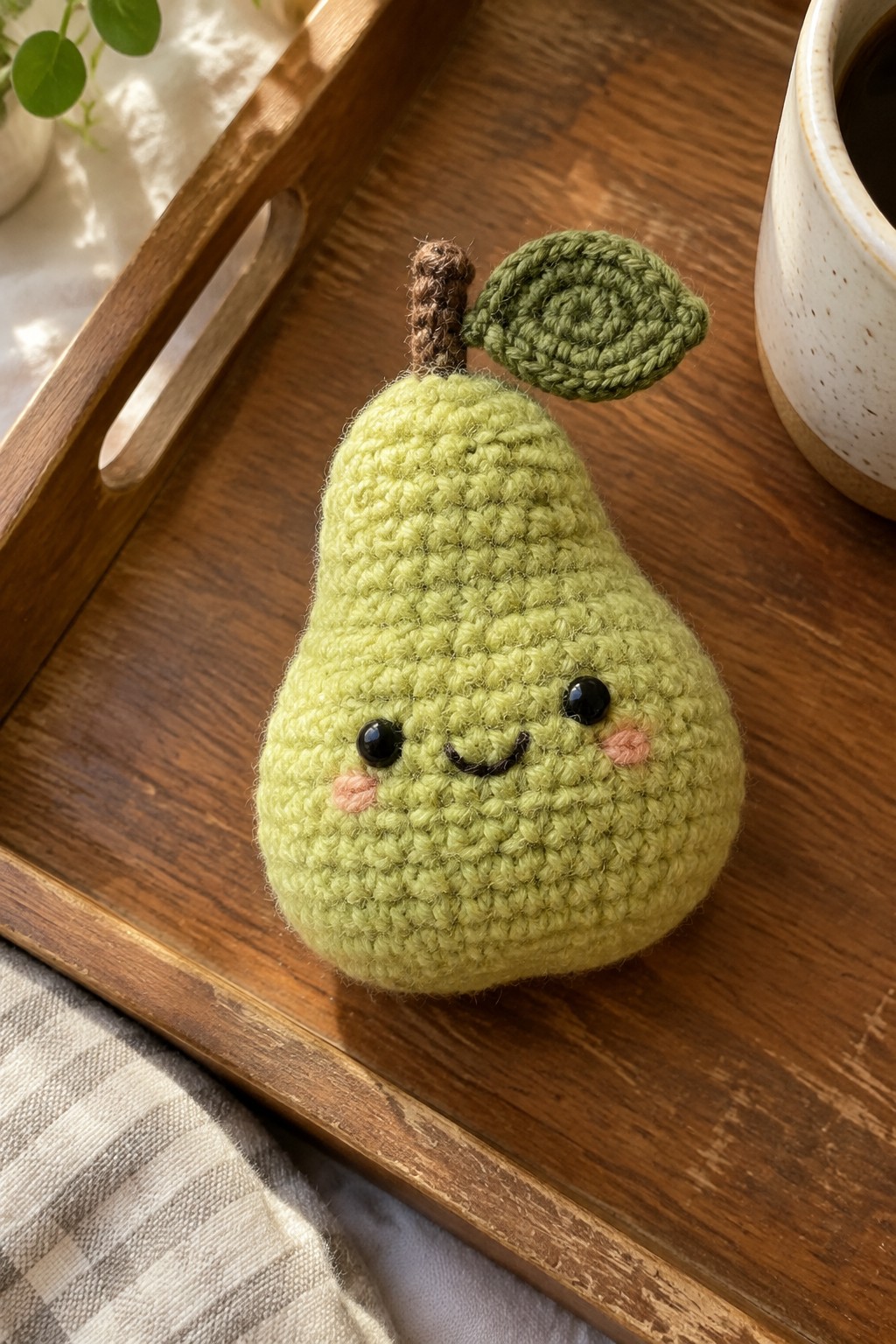 Crocheted green pear with smiling face on wooden tray beside coffee mug
