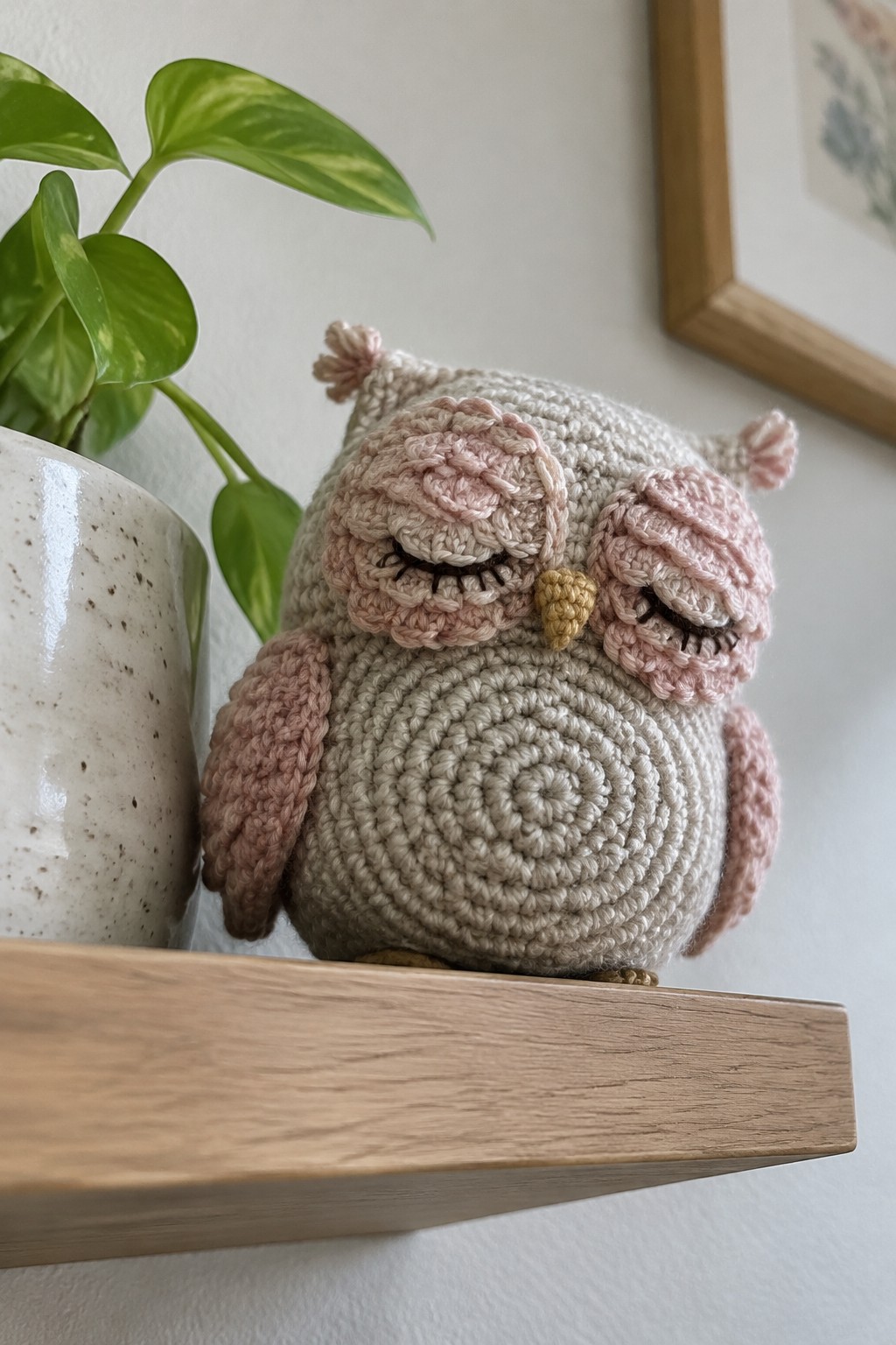 Crocheted sleeping owl in beige with pink accents on wooden shelf beside plant