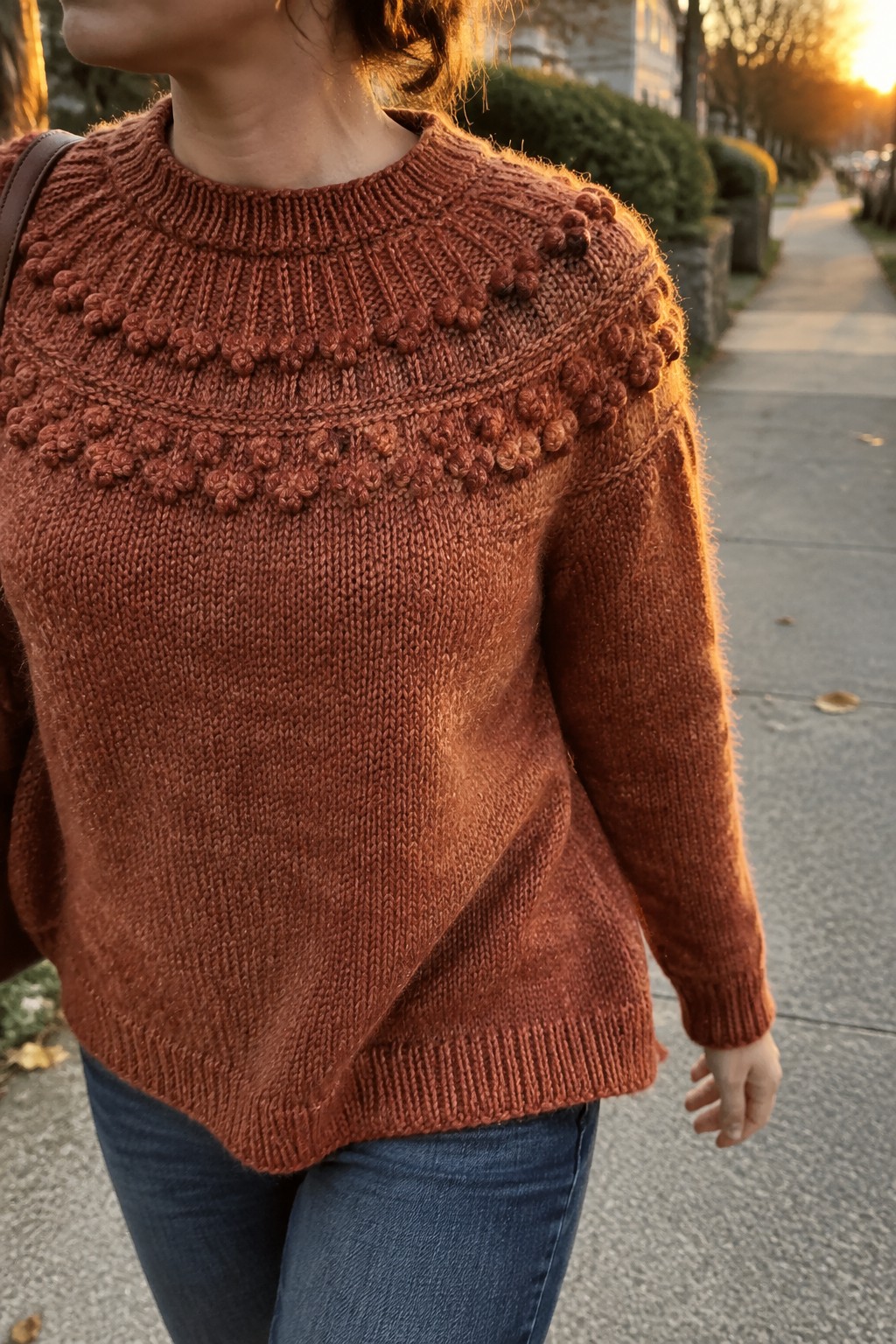 Rust-colored bobble-yoke knit sweater with blue jeans on a sidewalk at sunset