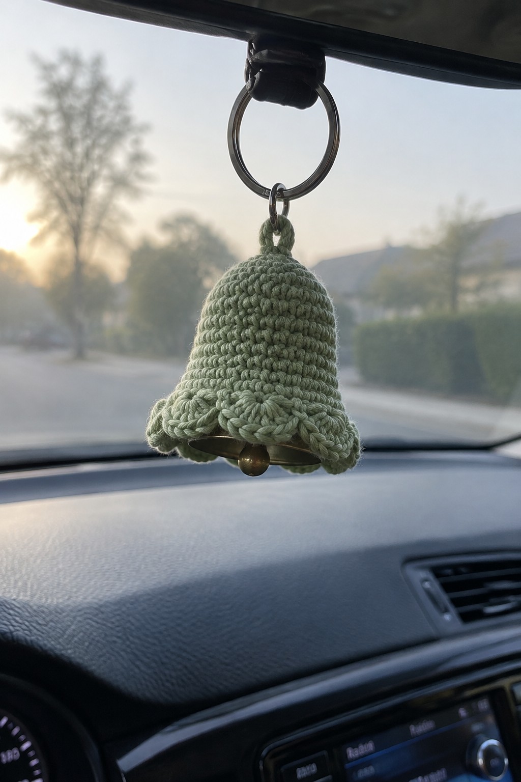 Sage green crocheted bell ornament dangling from car rearview mirror at sunset