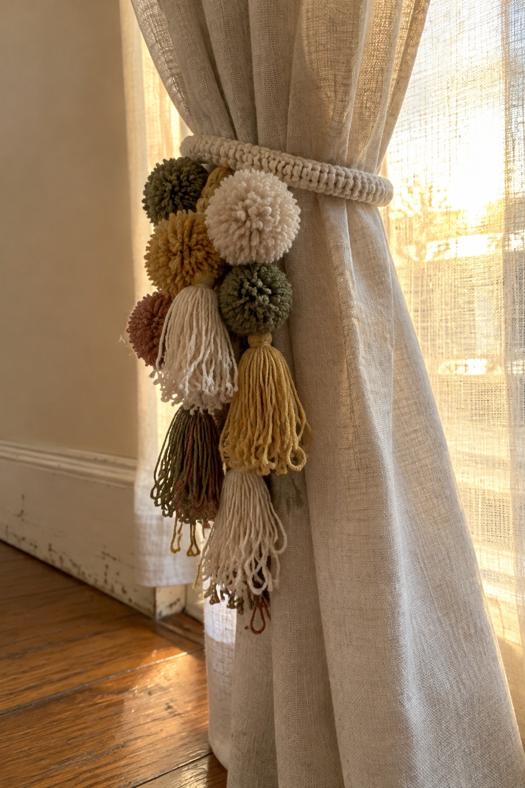 Beige linen curtain secured by crocheted tieback with colorful pom-poms and tassels.