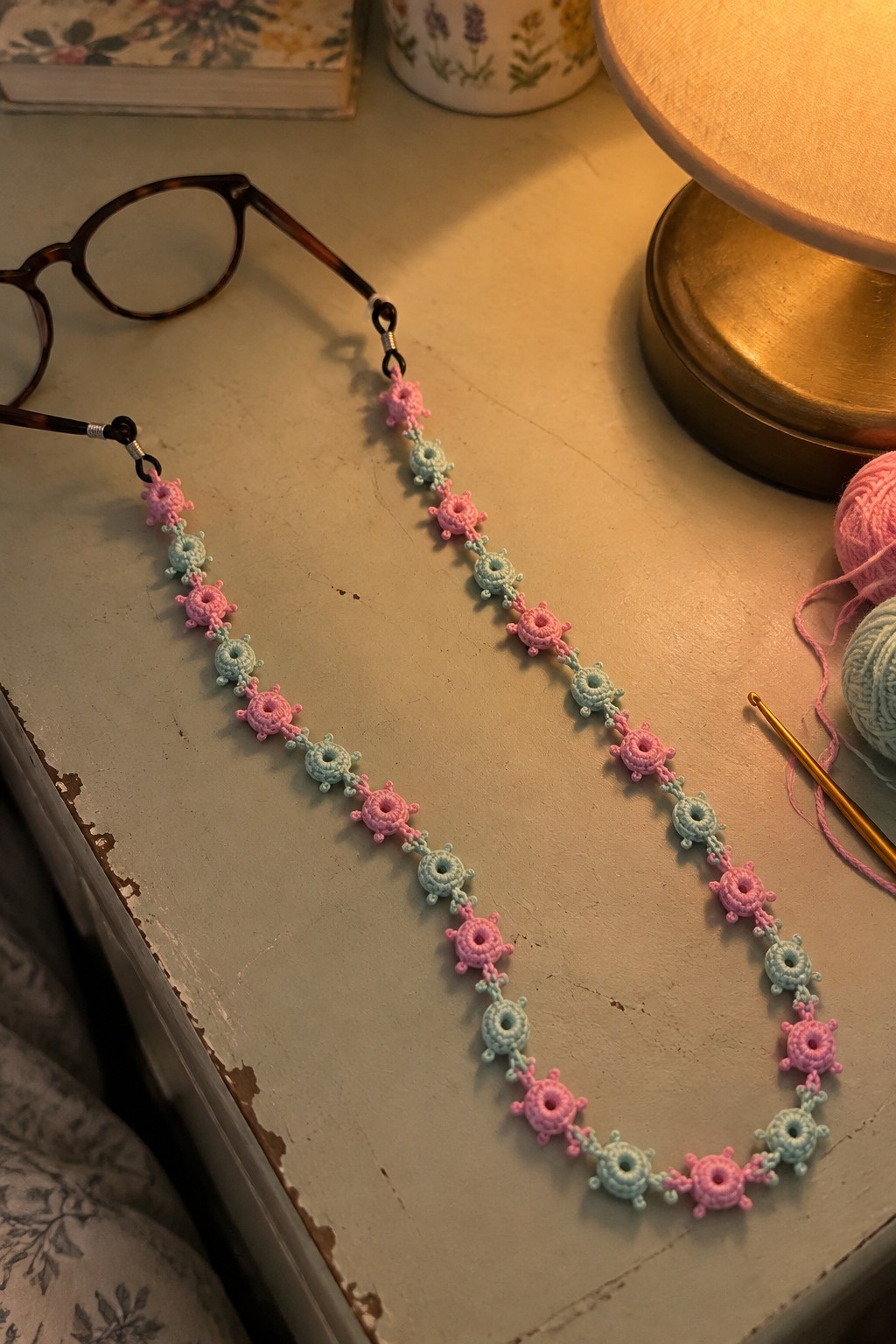 Pink and blue crocheted glasses chain on wooden table with yarn and lamp.