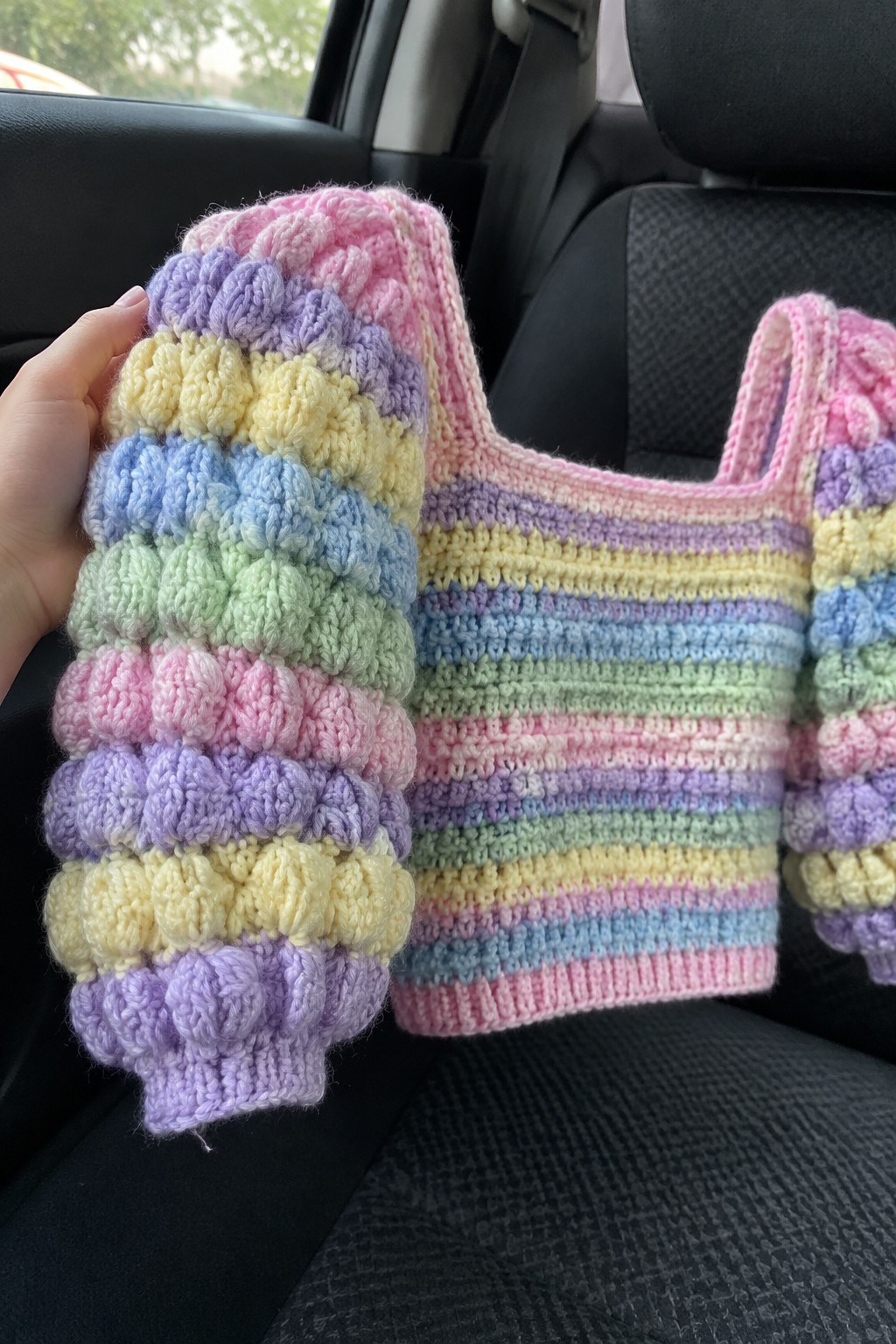 Hand holding rainbow crocheted top with bobble texture and stripes in car.