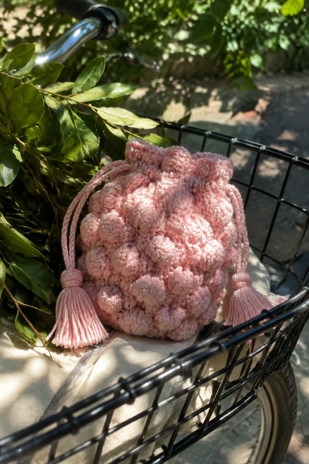 Pink bobble-knit pouch with tassels in black bicycle basket amid green leaves