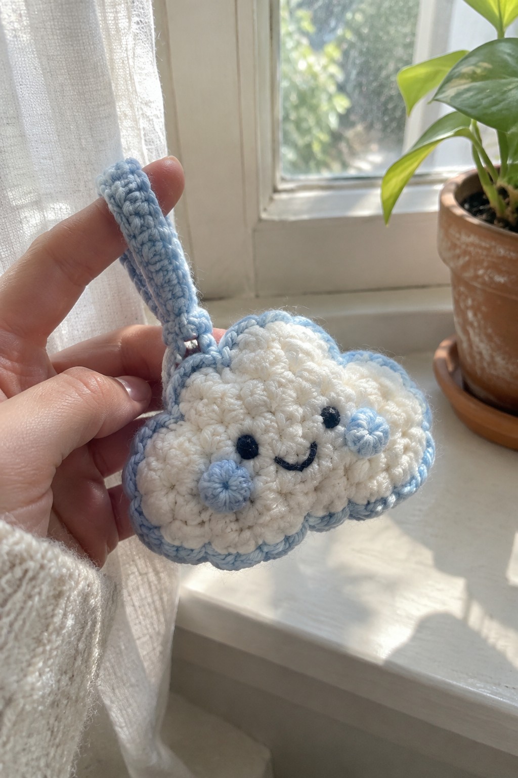 Hand holding white and blue crocheted cloud keychain with smiling face by window.