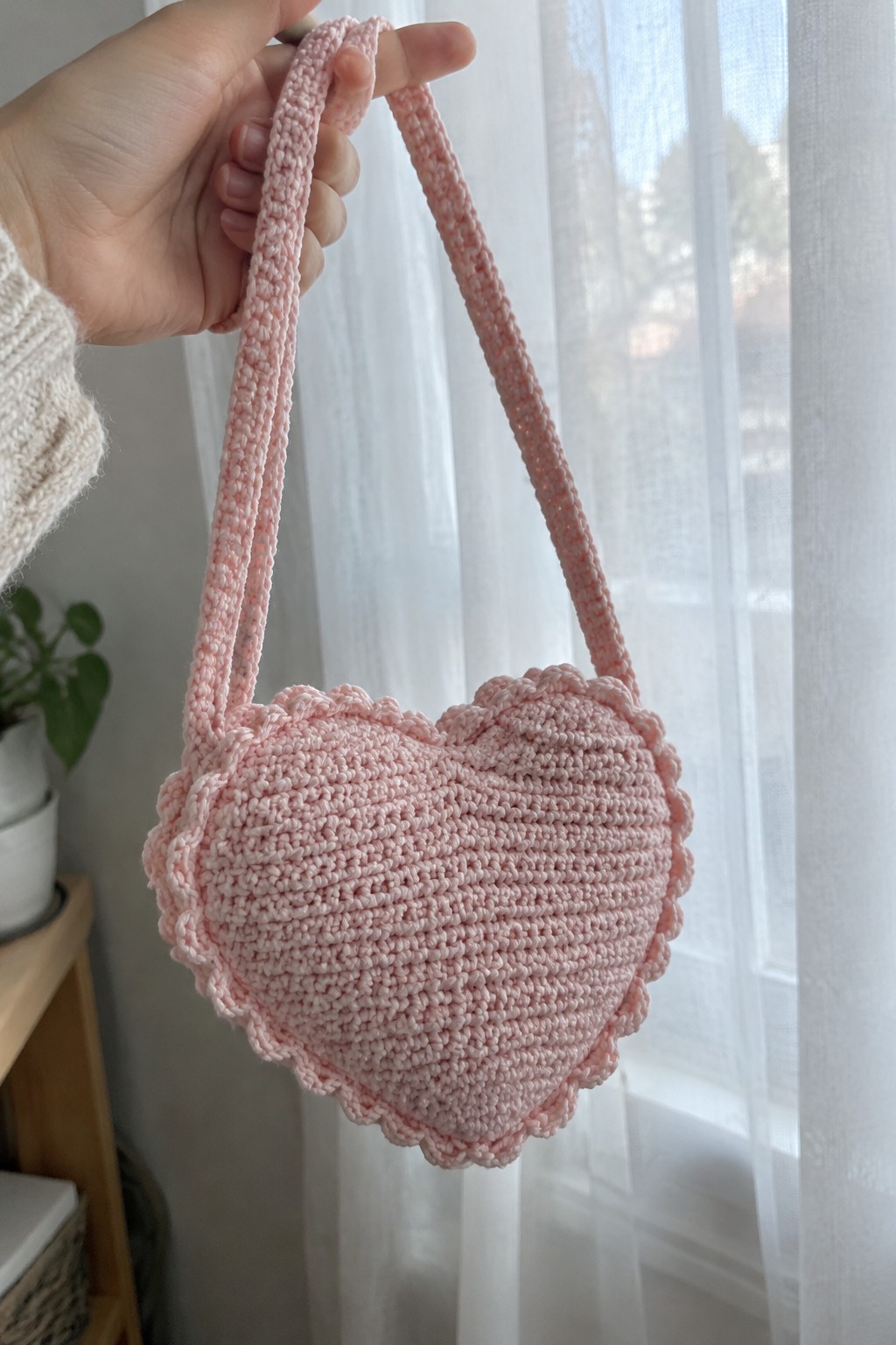 Hand in cream sweater holds pink crocheted heart-shaped bag with long strap.