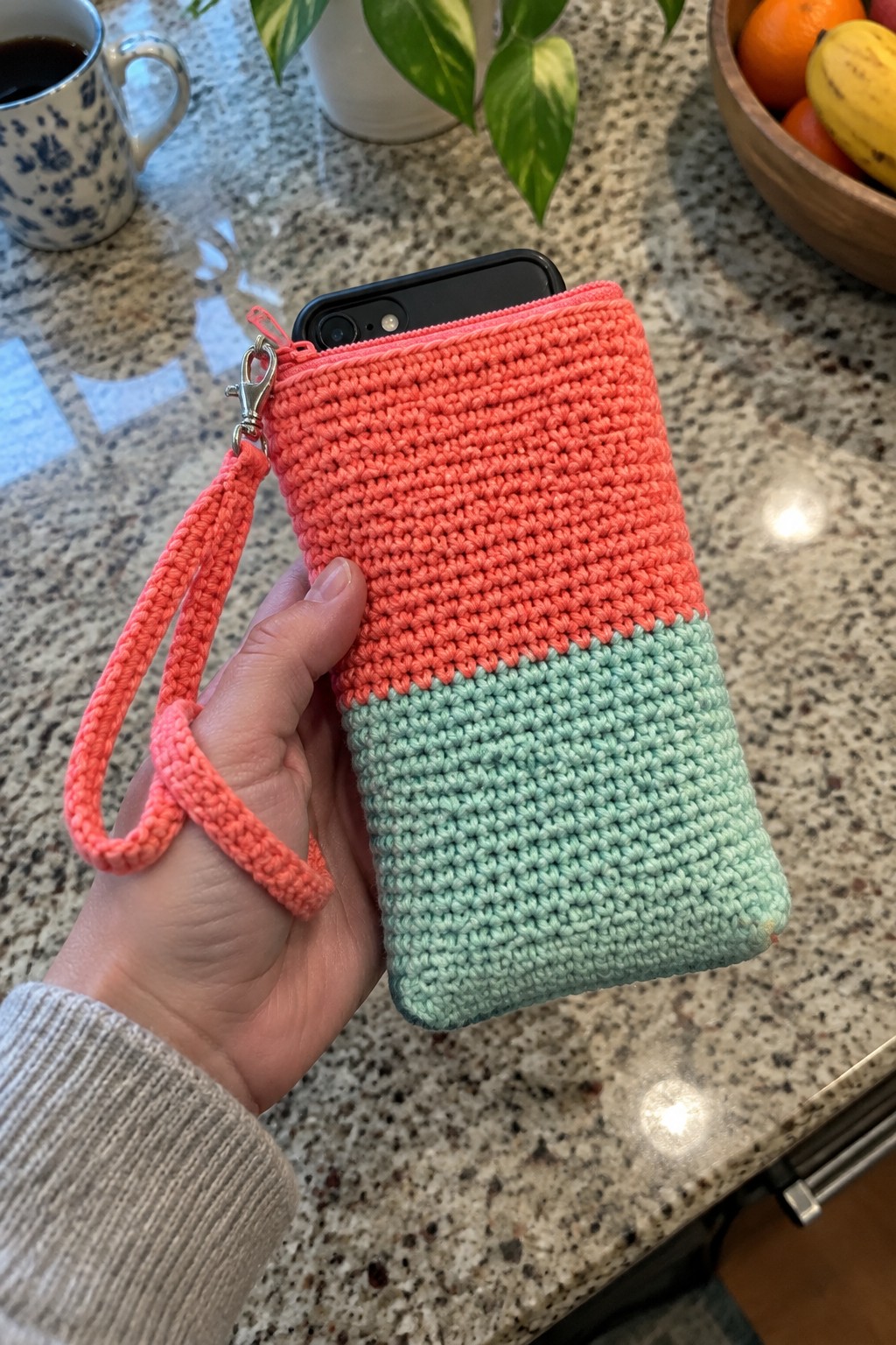 Hand holding coral and mint crocheted zippered pouch with smartphone and wrist strap