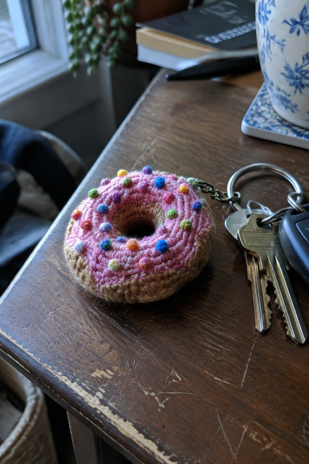 Crocheted pink donut keychain with colorful sprinkles beside keys on wooden table