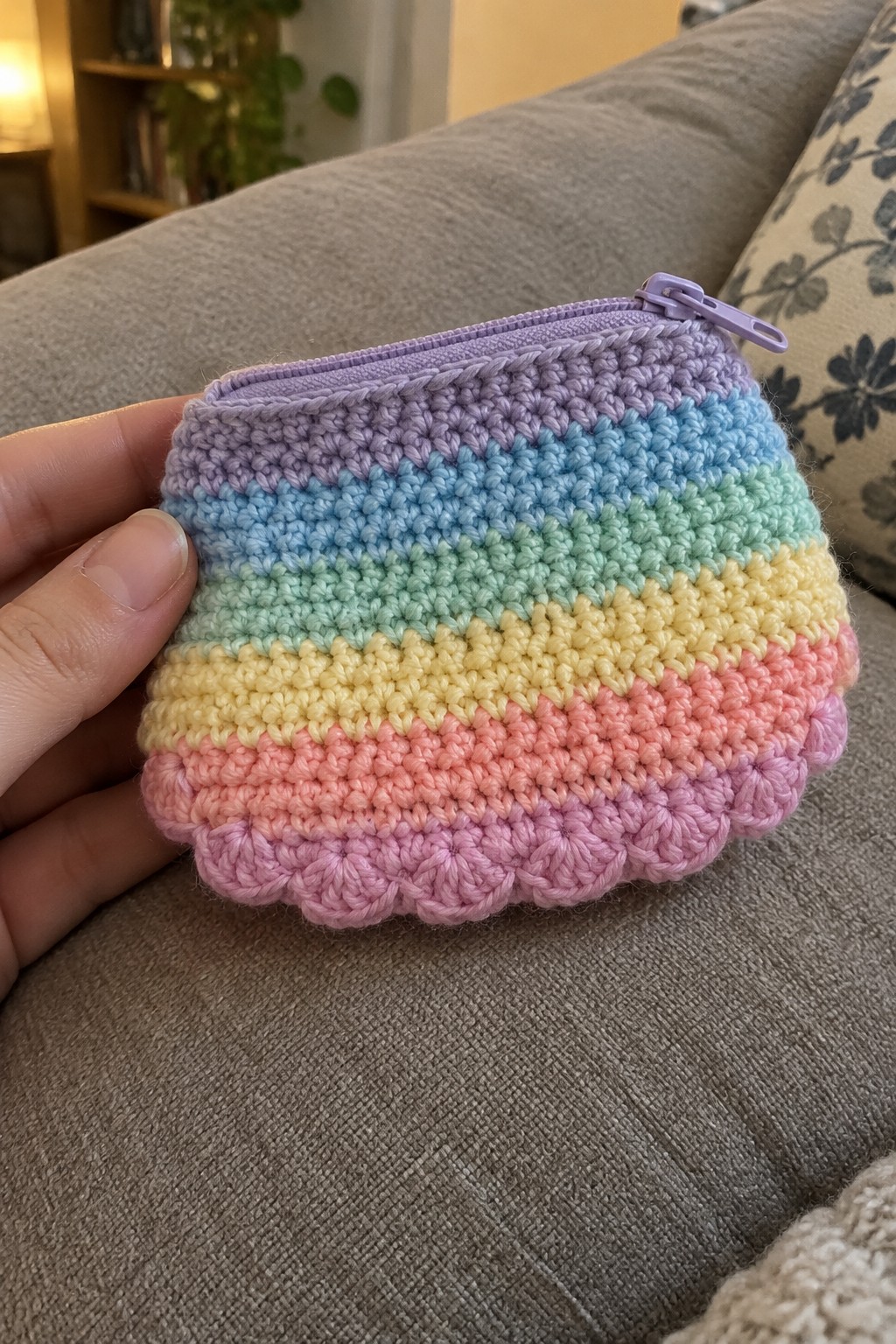 A hand holds a small crocheted pouch with horizontal rainbow stripes and a purple zipper against a gray fabric background.