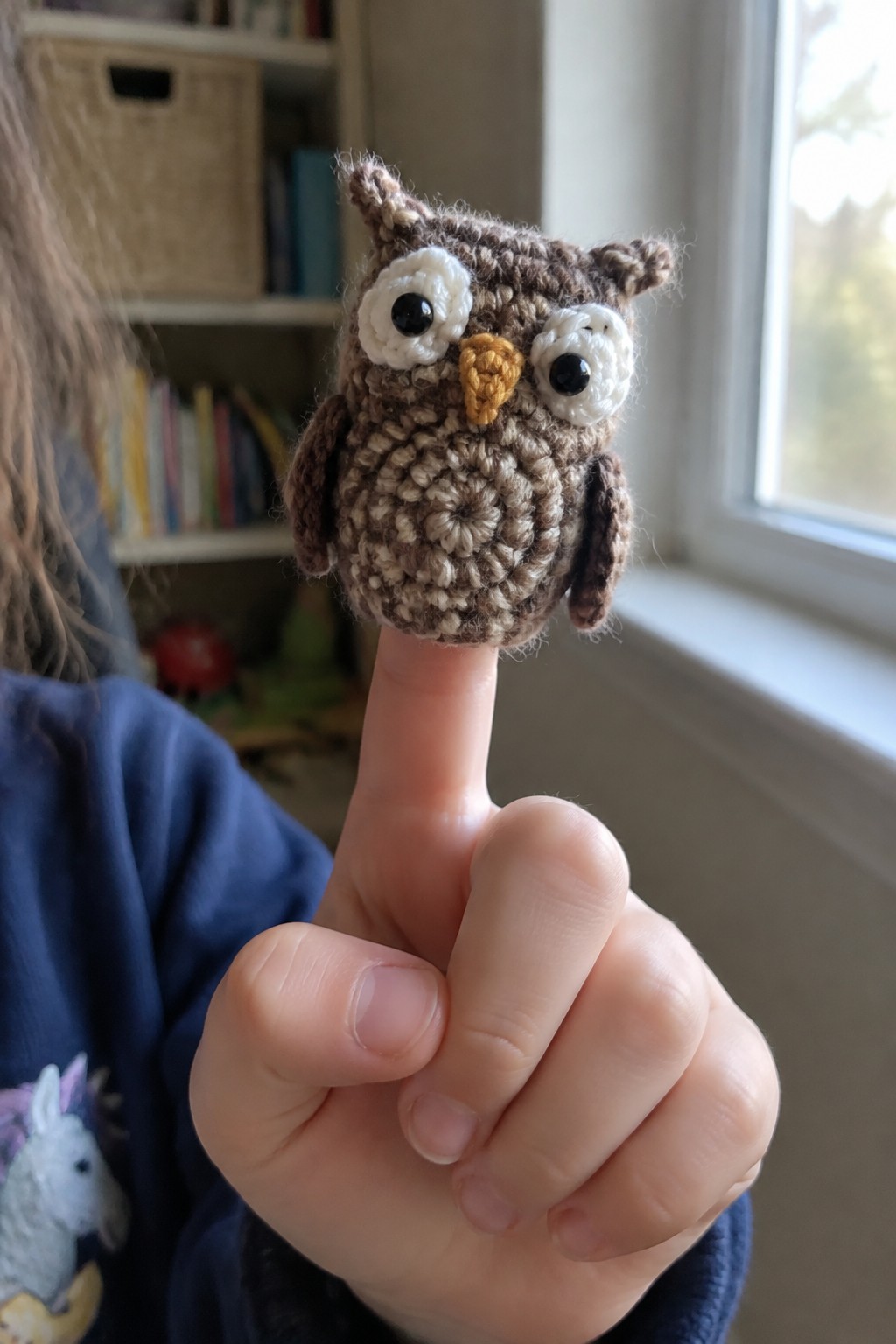 Crocheted brown owl finger puppet on thumb, blue unicorn shirt, bookshelf behind