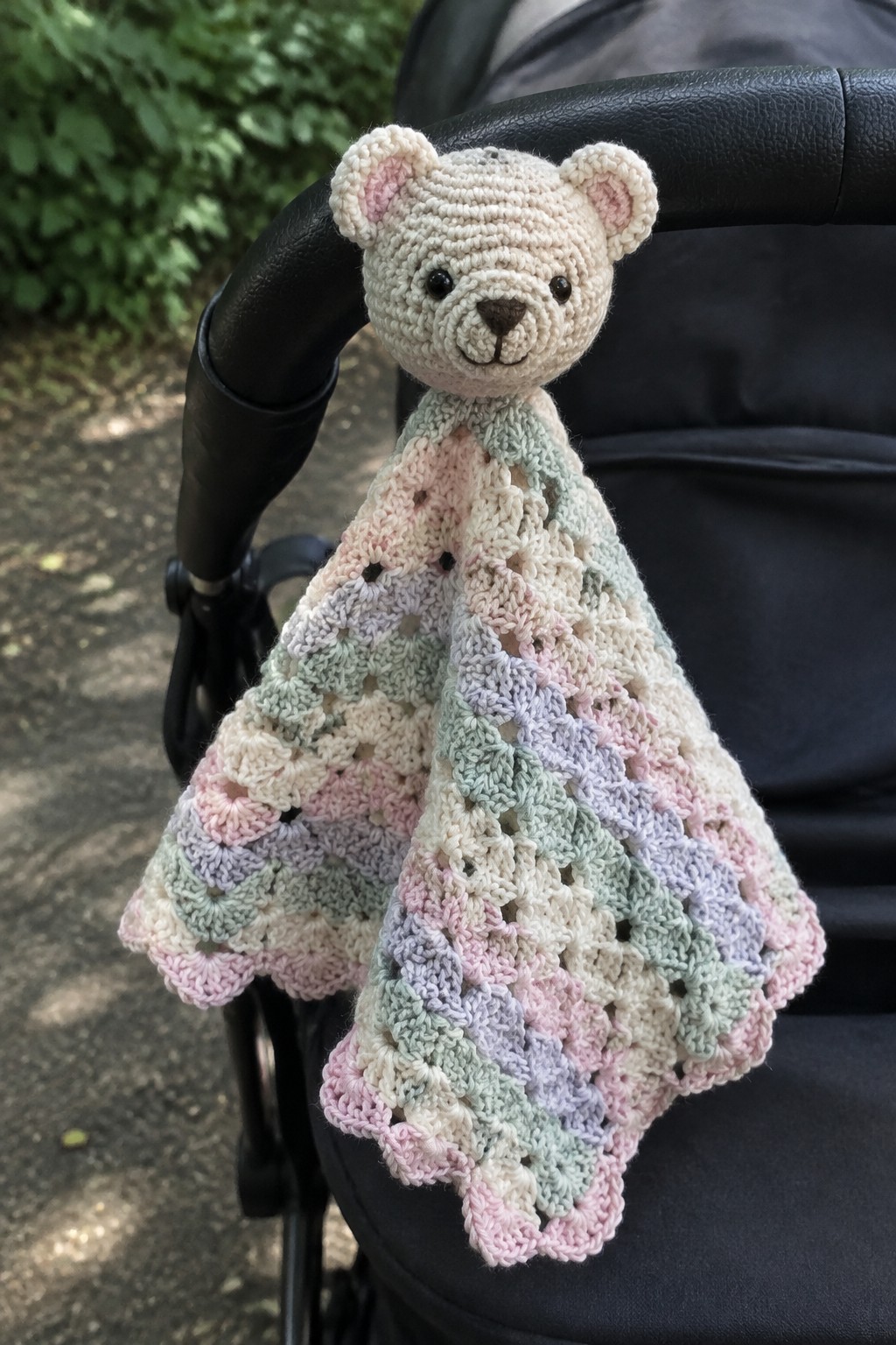 Crocheted teddy bear head on pastel lace blanket over black stroller handle.