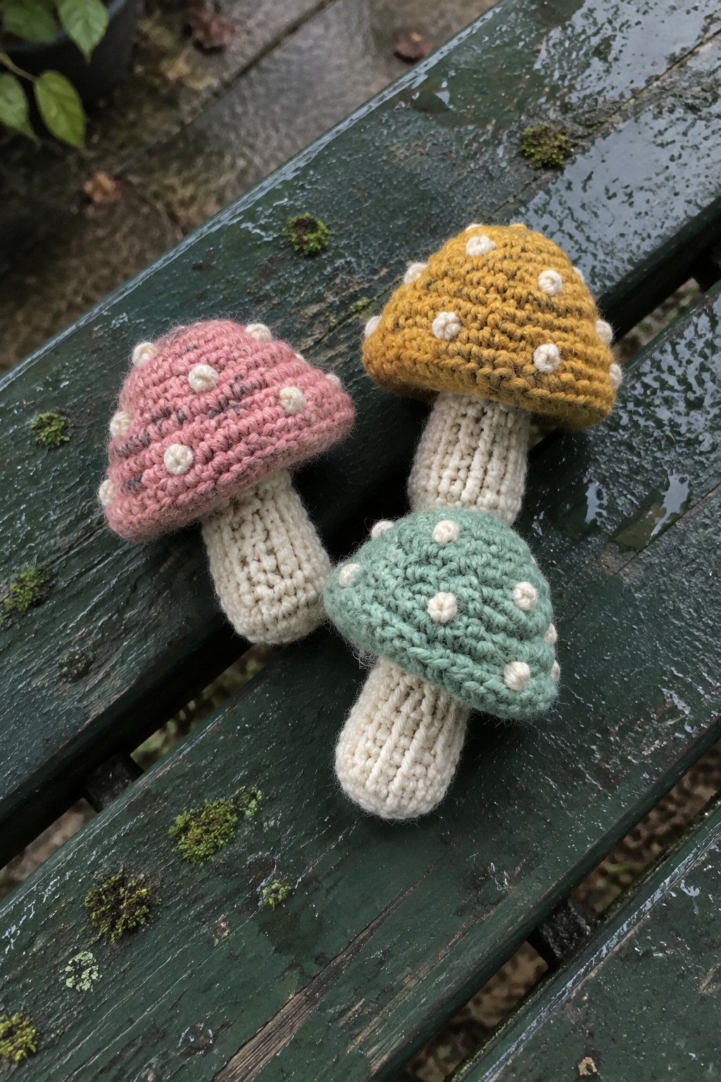 Three crocheted mushrooms with spotted caps on a wet green bench.