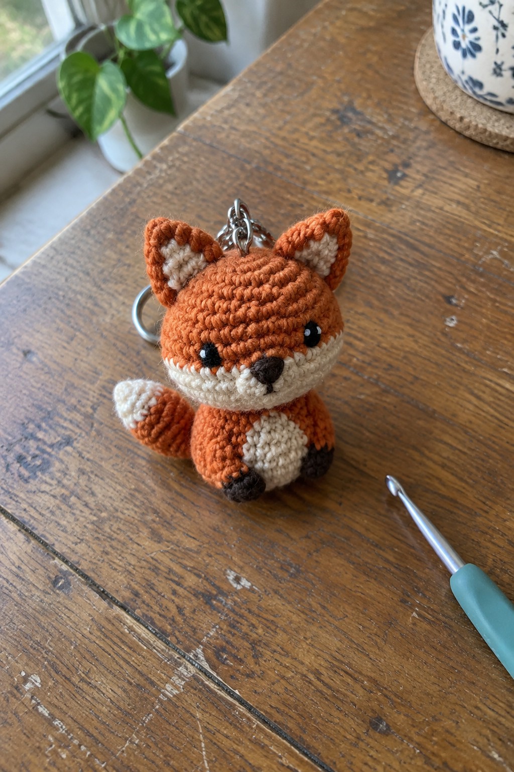 A small crocheted orange fox keychain with white and brown details rests on a wooden table beside a crochet hook.